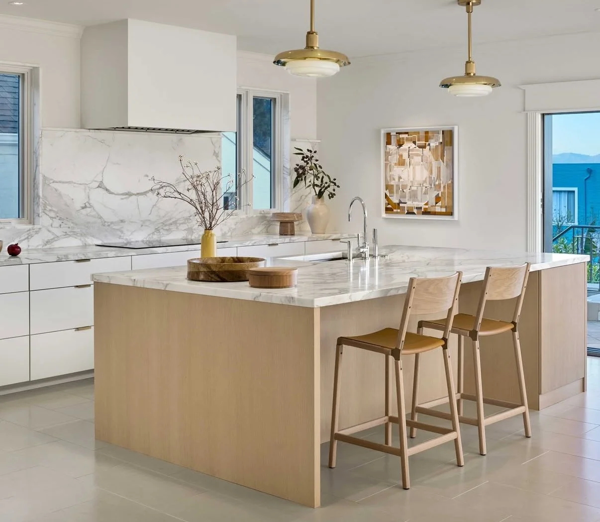 Modern kitchen with white walls, marble countertops, a large marble island, two wooden bar stools, and gold pendant lights.