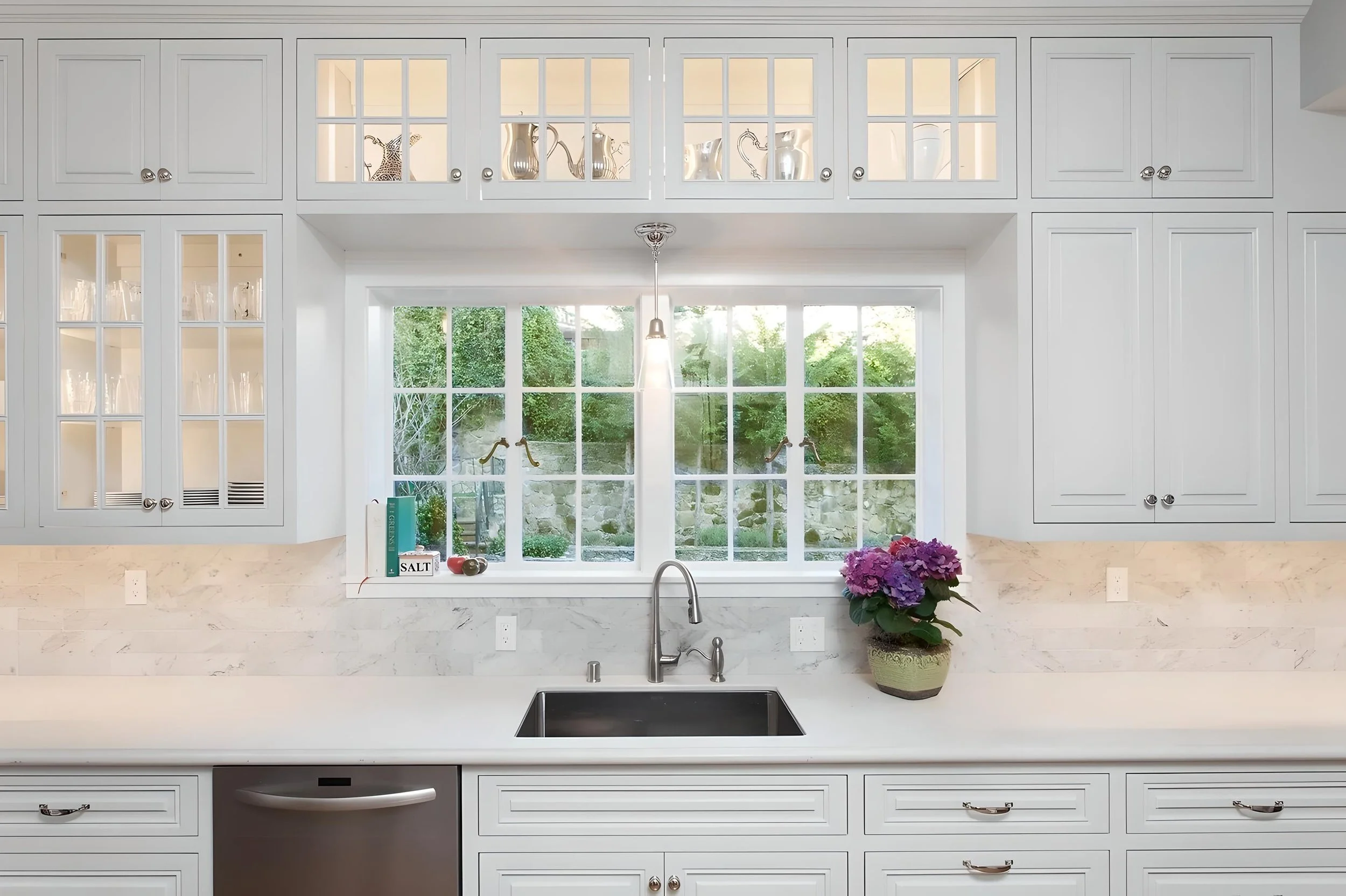 Kitchen with white cabinetry, a large window above the sink, and decorative glass-front cabinets. A potted purple hydrangea sits on the countertop beside a book and salt container. The window reveals a backyard with greenery and stone wall.