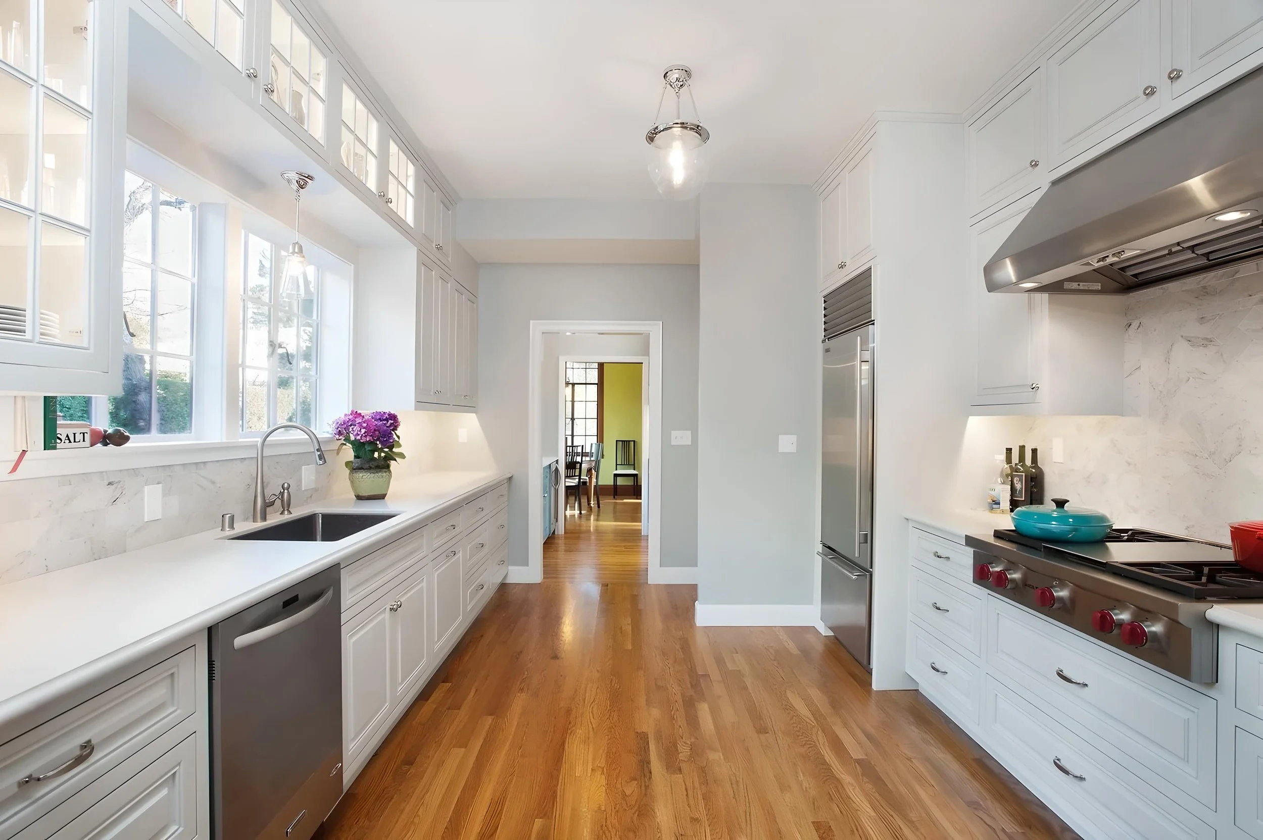 Modern kitchen with white cabinets, a large window with a view outside, a kitchen sink with a potted plant, wooden floor, and stainless steel appliances including a stove and refrigerator.