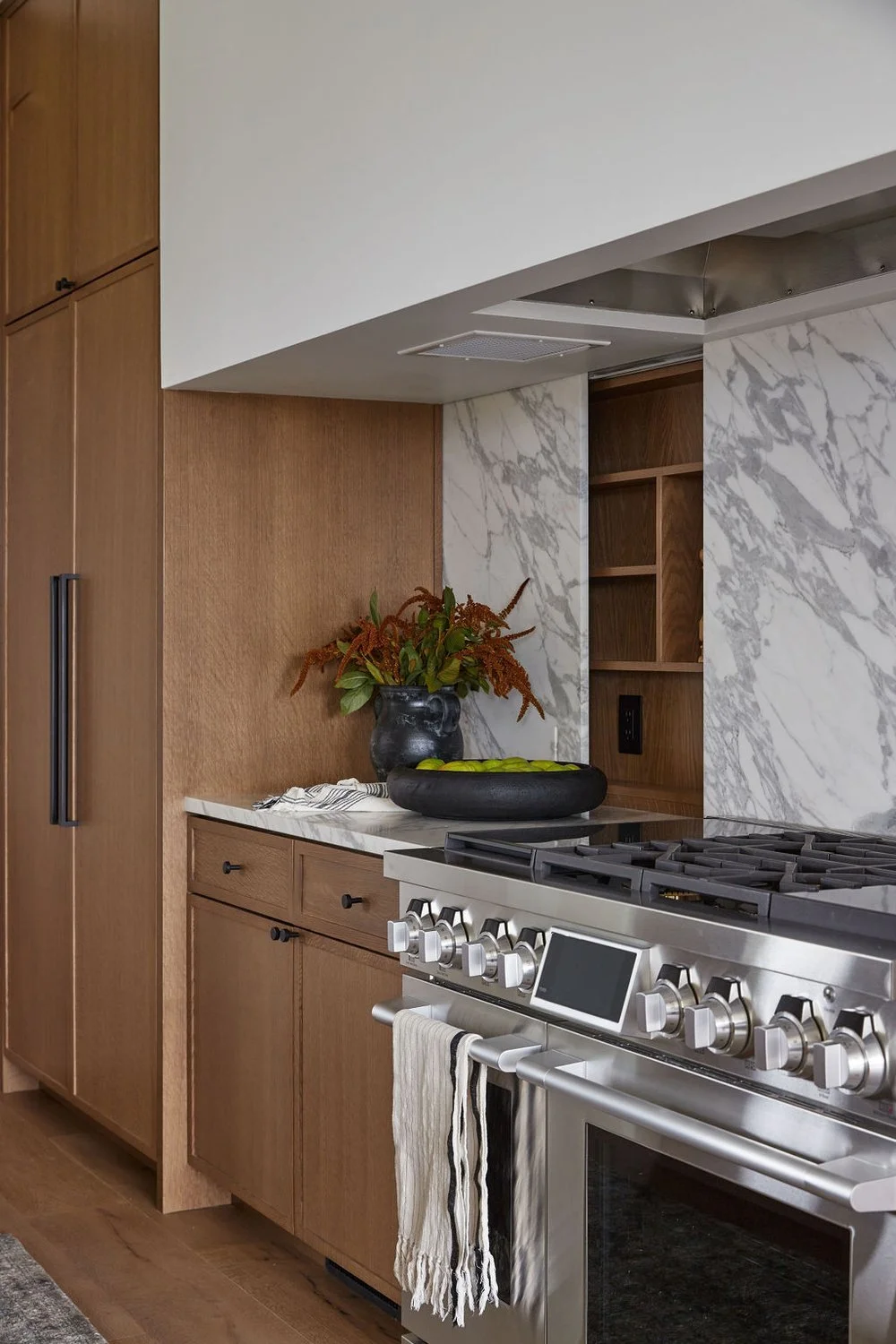 A modern kitchen corner with wooden cabinets, a white marble countertop, a black vase with plant, a black bowl with green apples, and a stainless steel stove with control knobs and a digital display.
