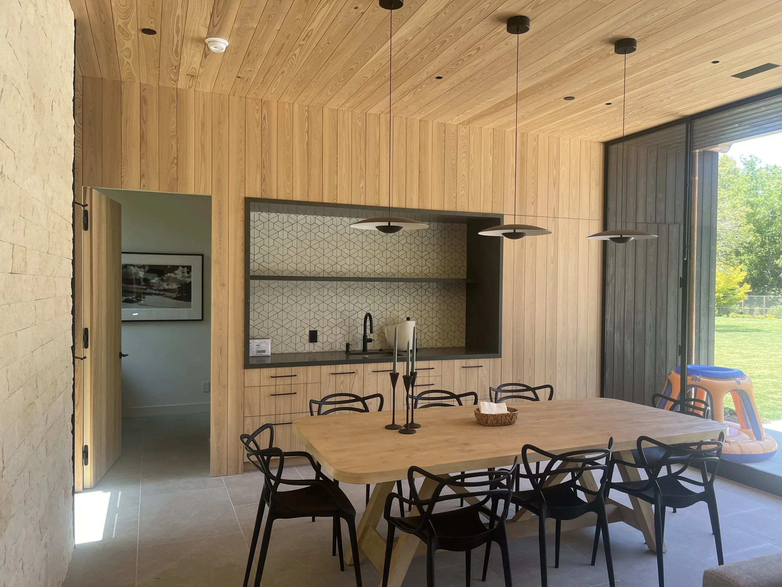 Modern dining room interior with wooden walls and ceiling, geometric backsplash, large wooden table with black chairs, pendant lighting, and a view of a backyard through glass doors.