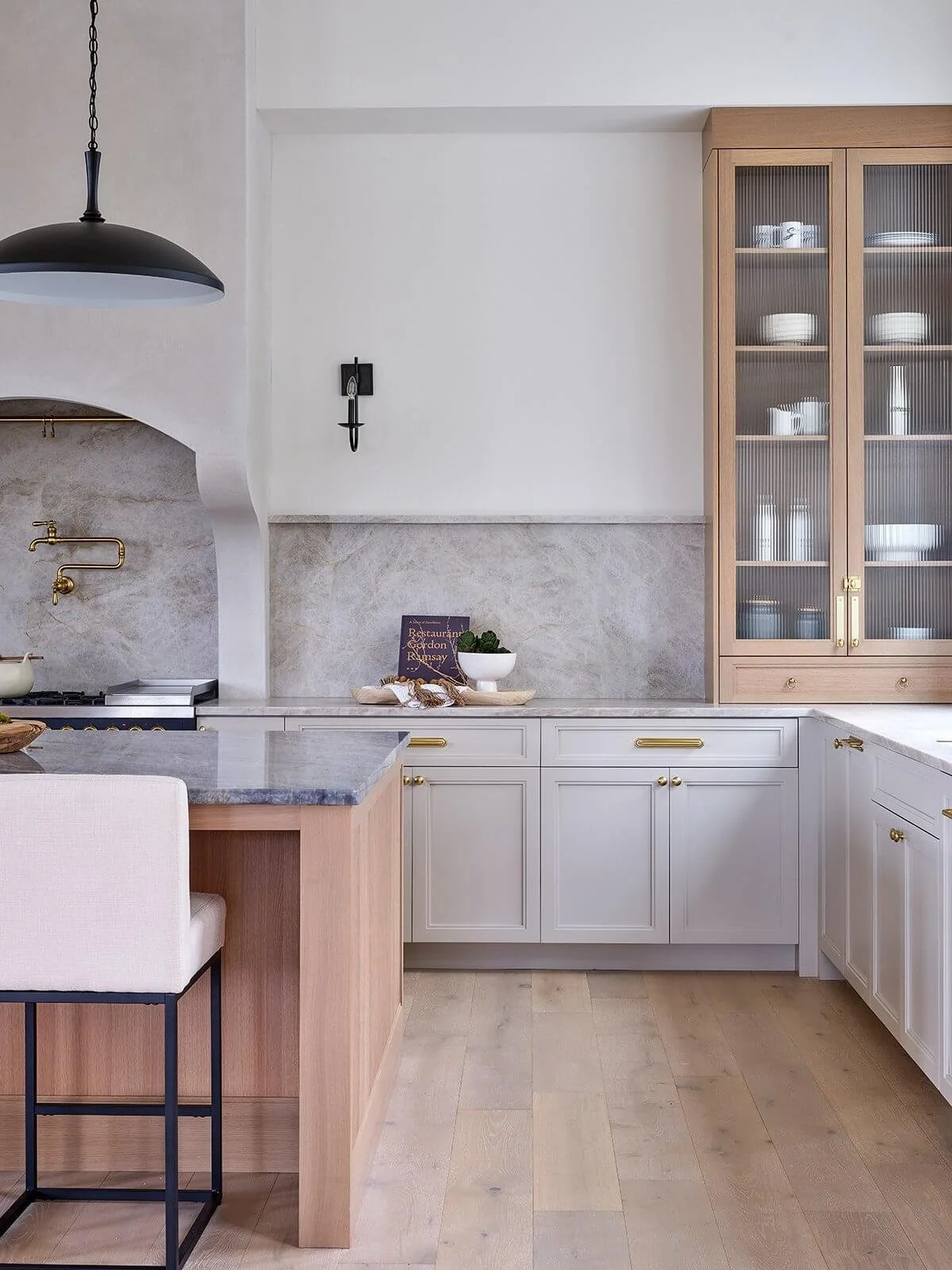 A modern kitchen with beige and white cabinetry, a gray marble countertop, a glass-front cabinet with white dishes, and a black pendant light.