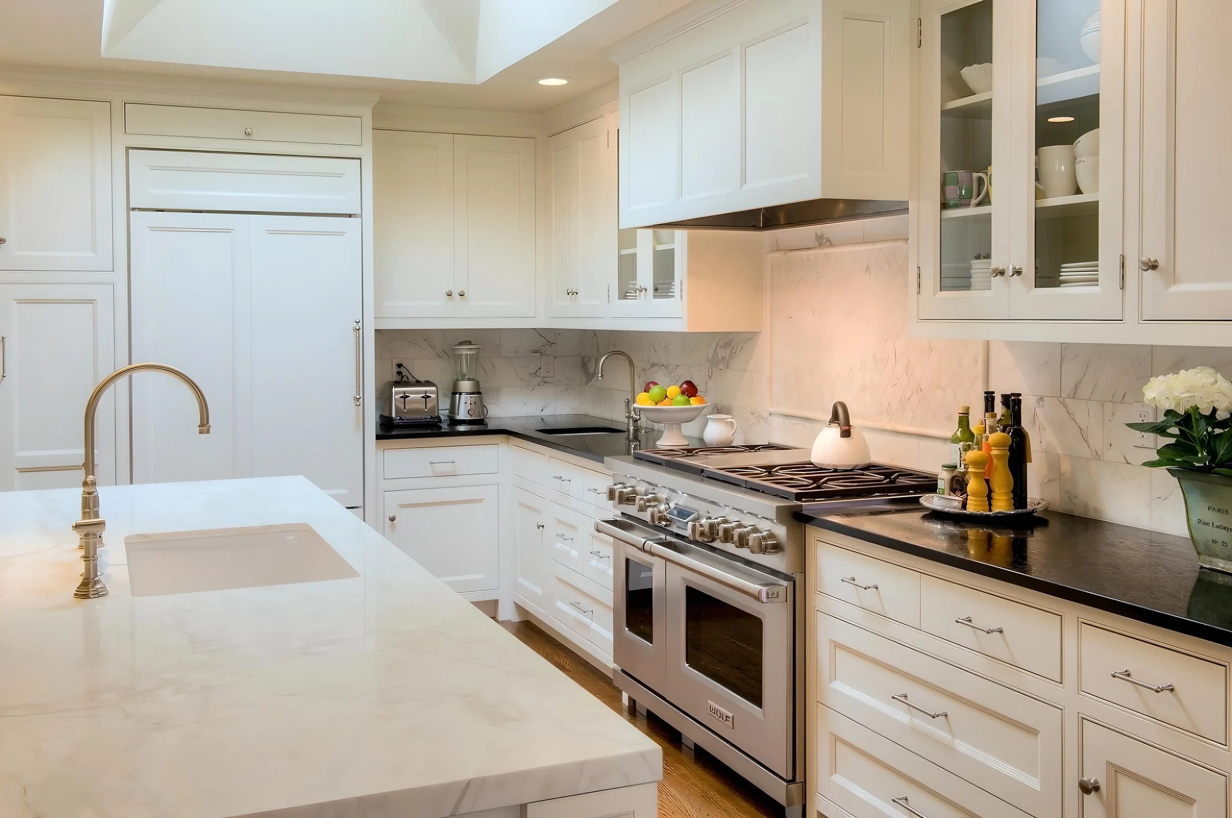 Modern white kitchen with marble countertops, stainless steel appliances, and decorative items like fruit, flowers, and condiments.
