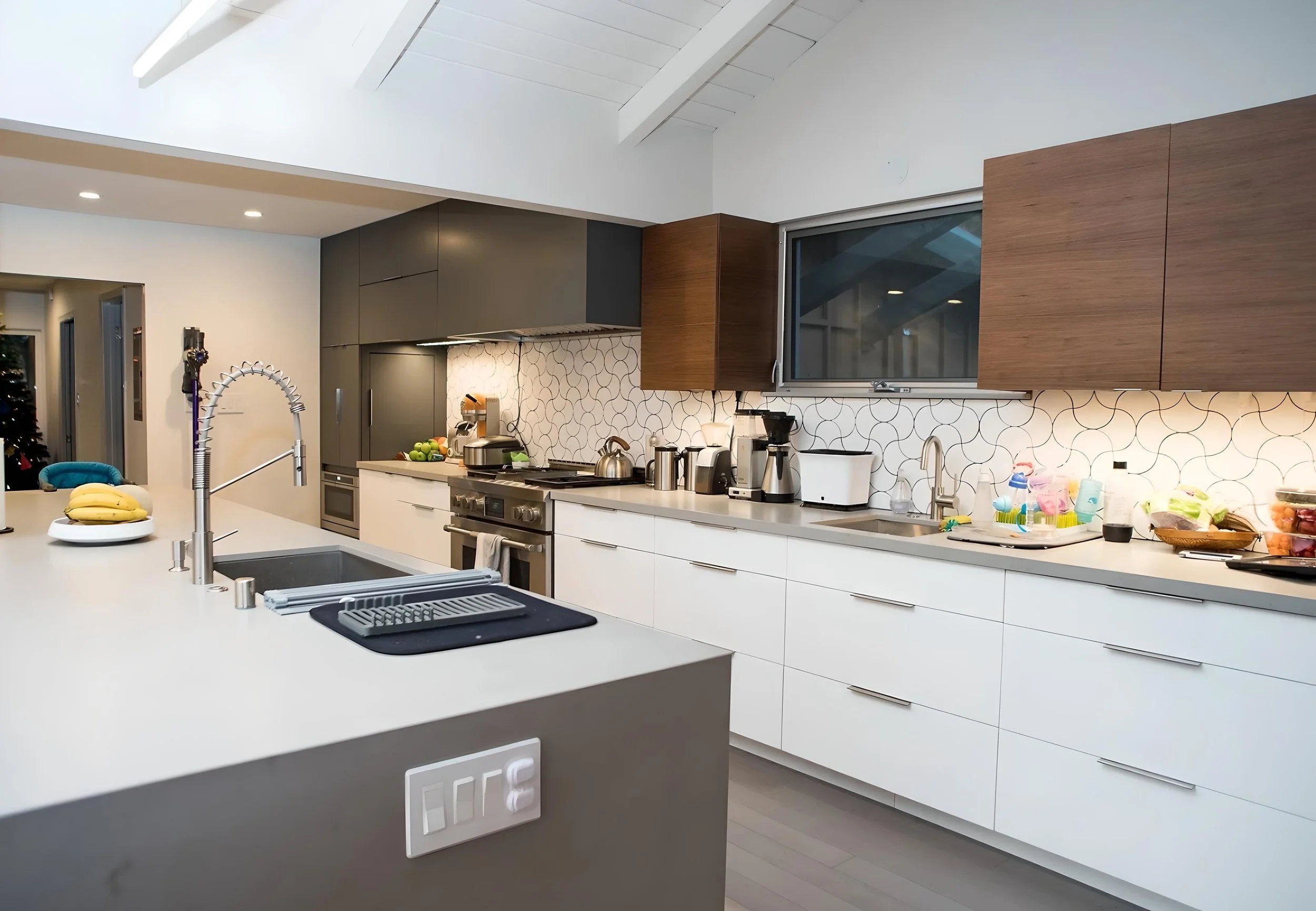 Modern kitchen with white counter, stainless steel appliances, dark gray and wooden cabinets, geometric tile backsplash, and various small appliances.