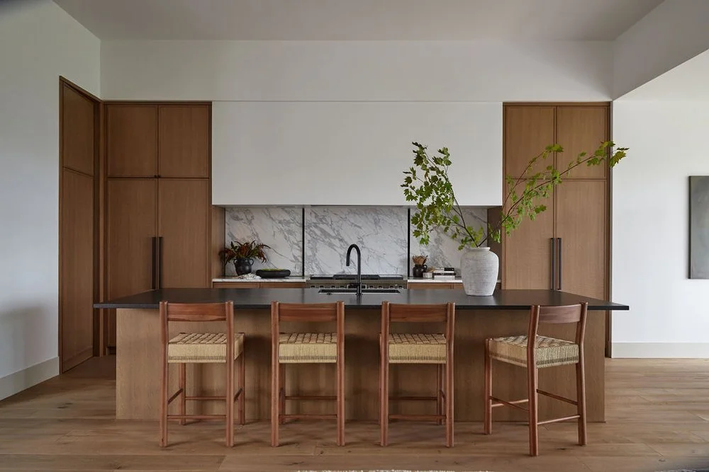 Modern kitchen with wooden cabinets, a black countertop, a marble backsplash, a black sink, five wooden chairs with woven seats around a large kitchen island, and a white vase with green branches.