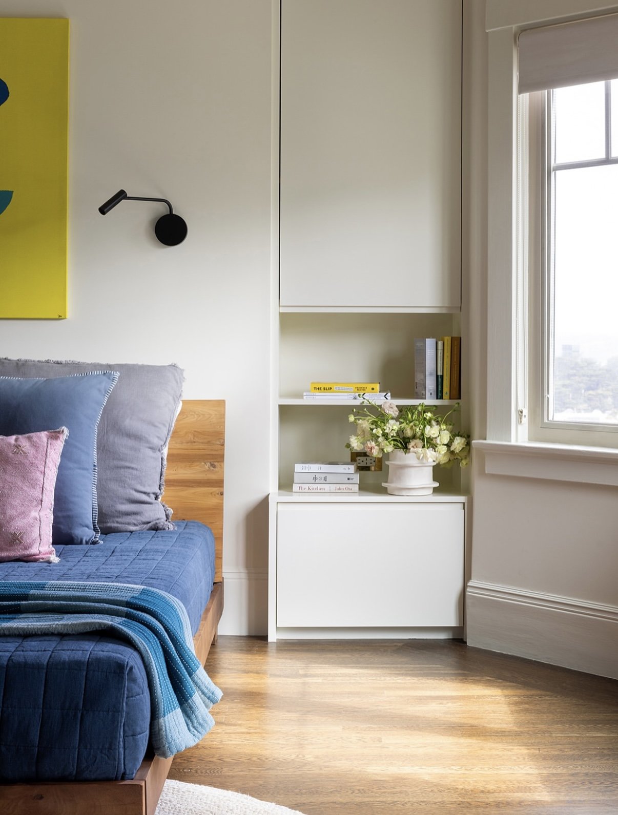 A bedroom corner with a bed having blue and purple pillows, a wooden headboard, and a blue quilt, alongside a white built-in shelving unit with books and a vase of white flowers, near a large window with daylight.