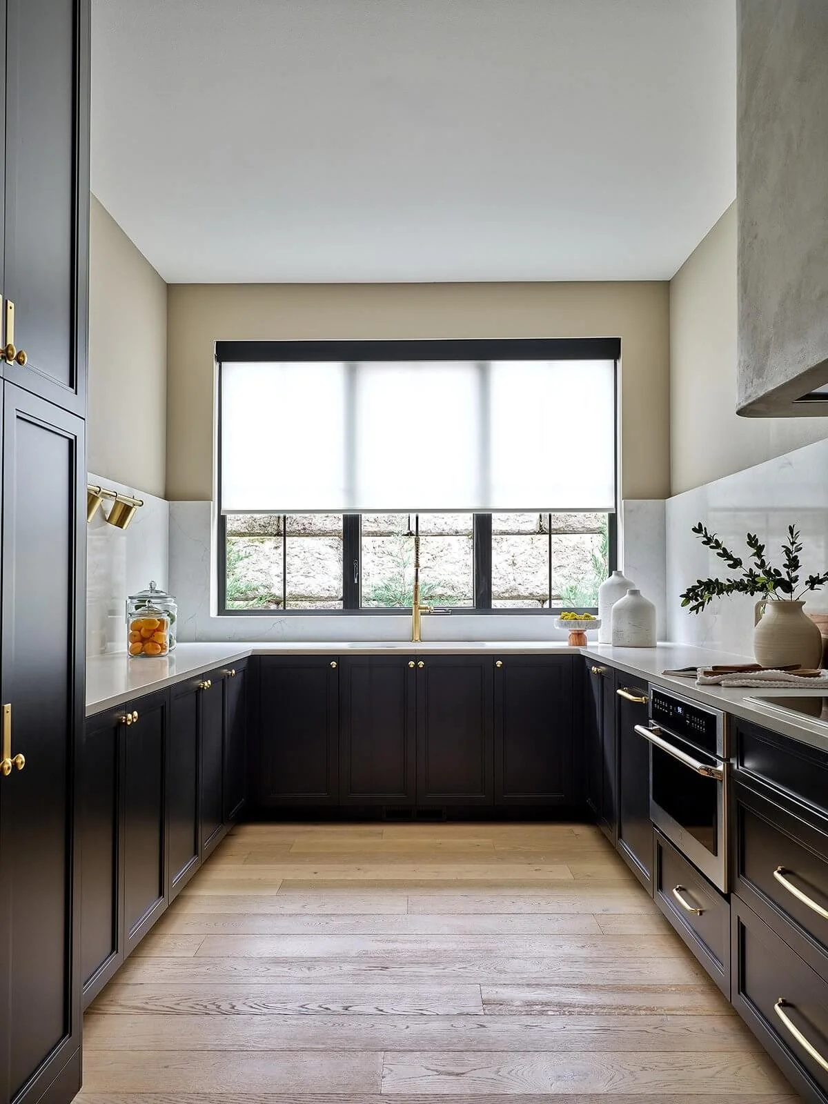 Modern kitchen with black cabinets, white countertops, a large window with a roller shade, and light wood flooring.