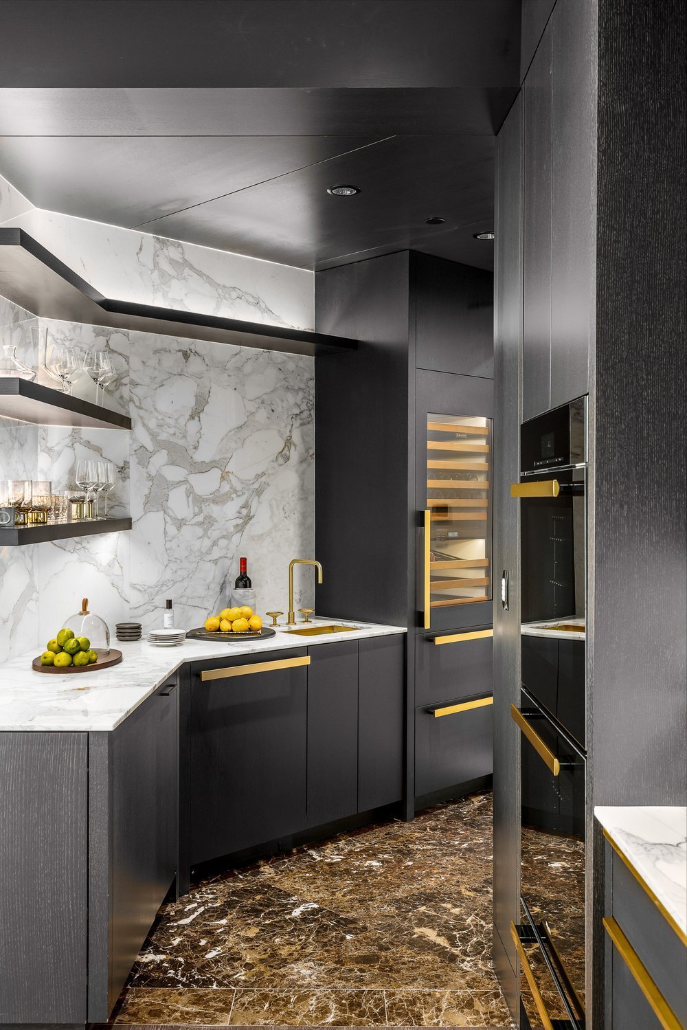 Modern kitchen with black cabinets, gold accents, marble countertops and backsplash, open shelves with glassware, a bottle of wine, and a bowl of lemons and limes.