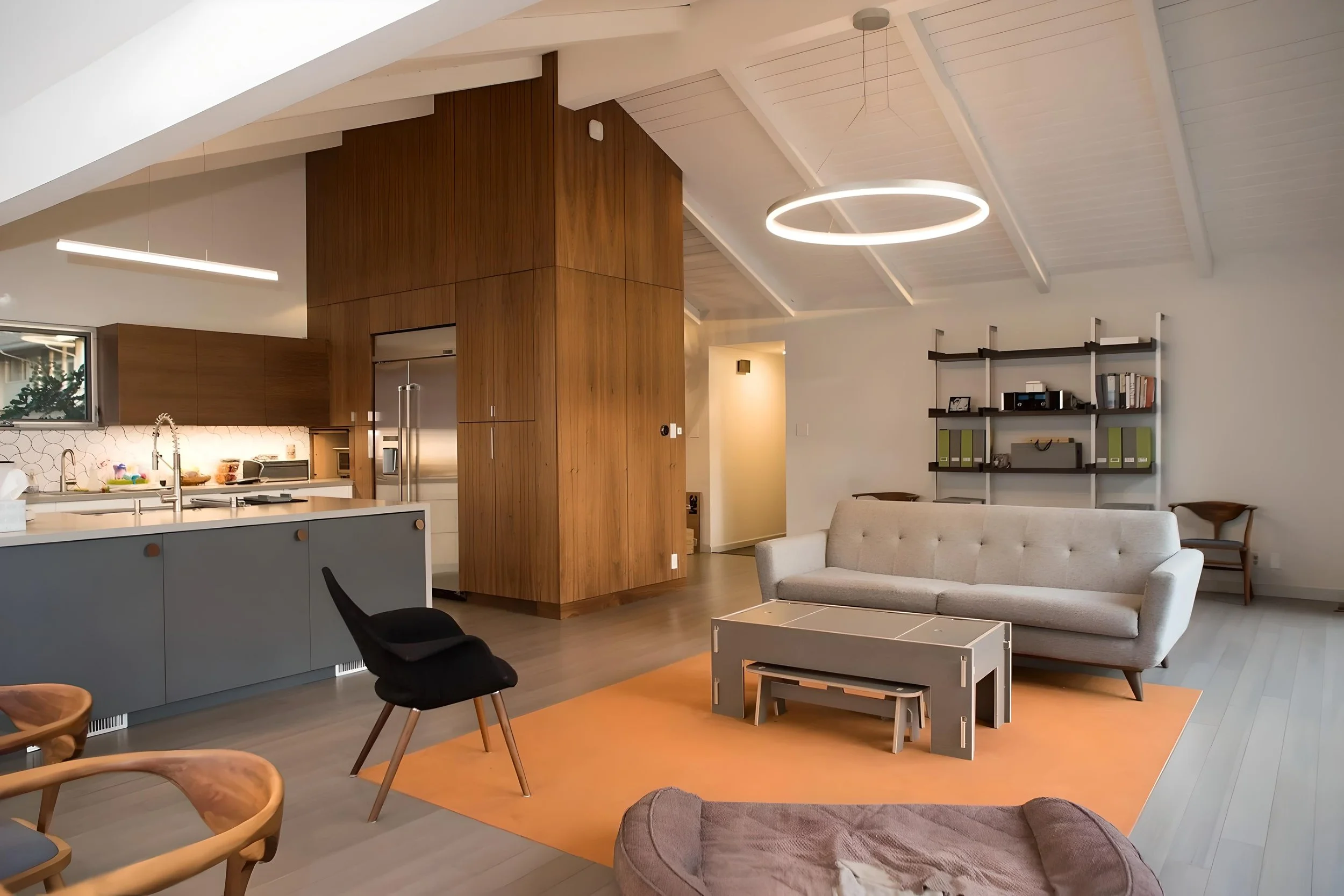 Open concept living room and kitchen with modern furniture, a white sofa, a black and wood chair, a gray table on an orange rug, wooden shelving with books and decorations, and a circular ceiling light.