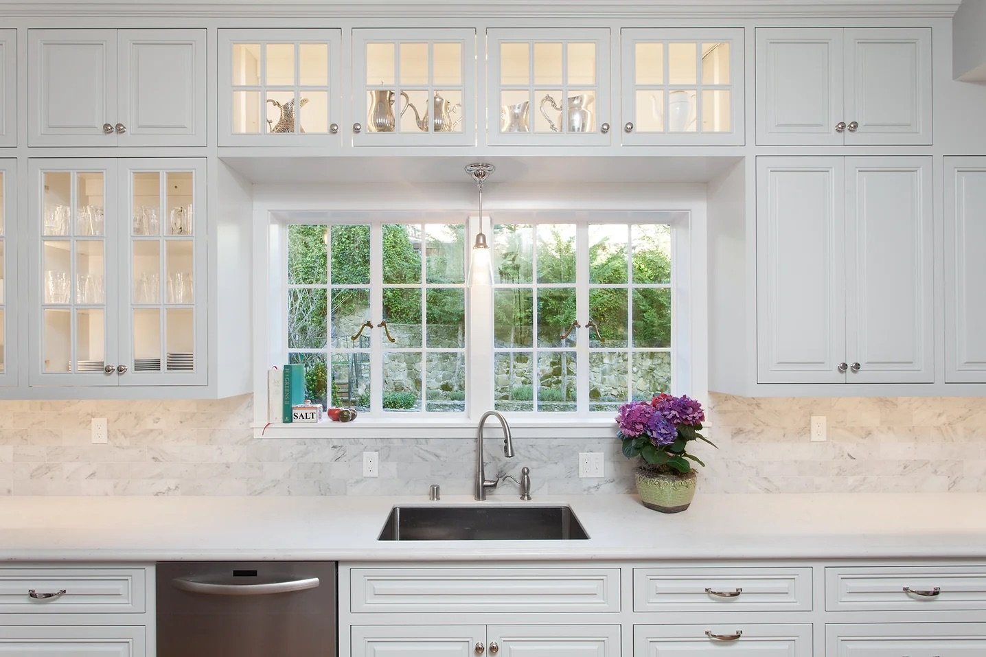 Modern kitchen with white cabinets, window, stainless steel sink, and countertops. Decorative items and flowers are placed around.