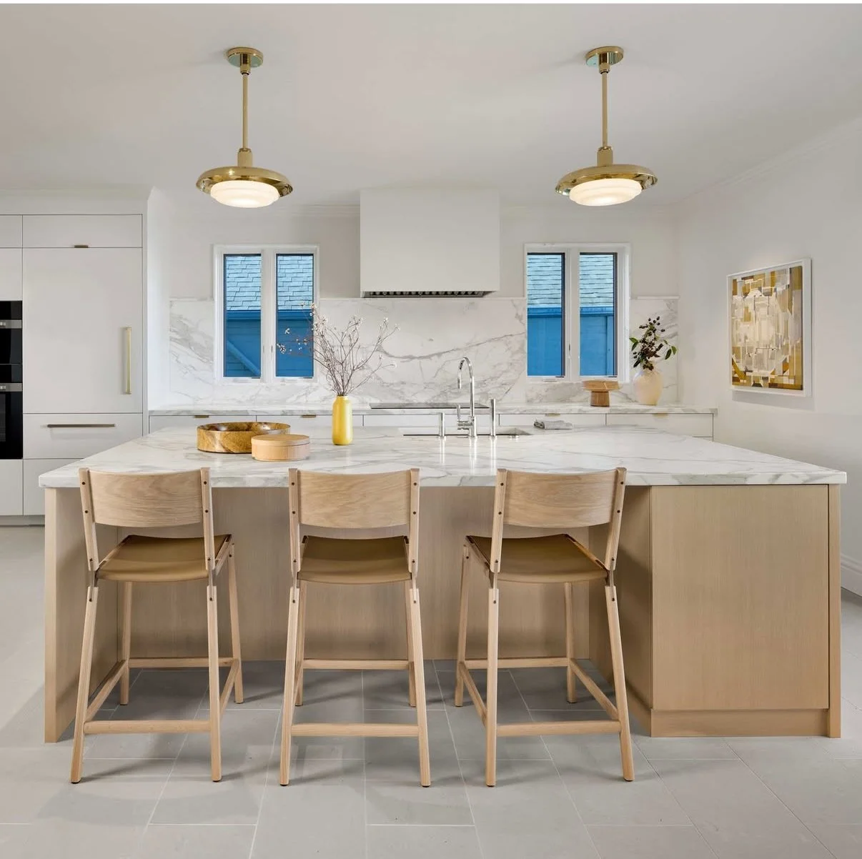 Modern kitchen with white marble island, light wood bar stools, and minimal decor, featuring white cabinetry, dual windows, and pendant lighting.