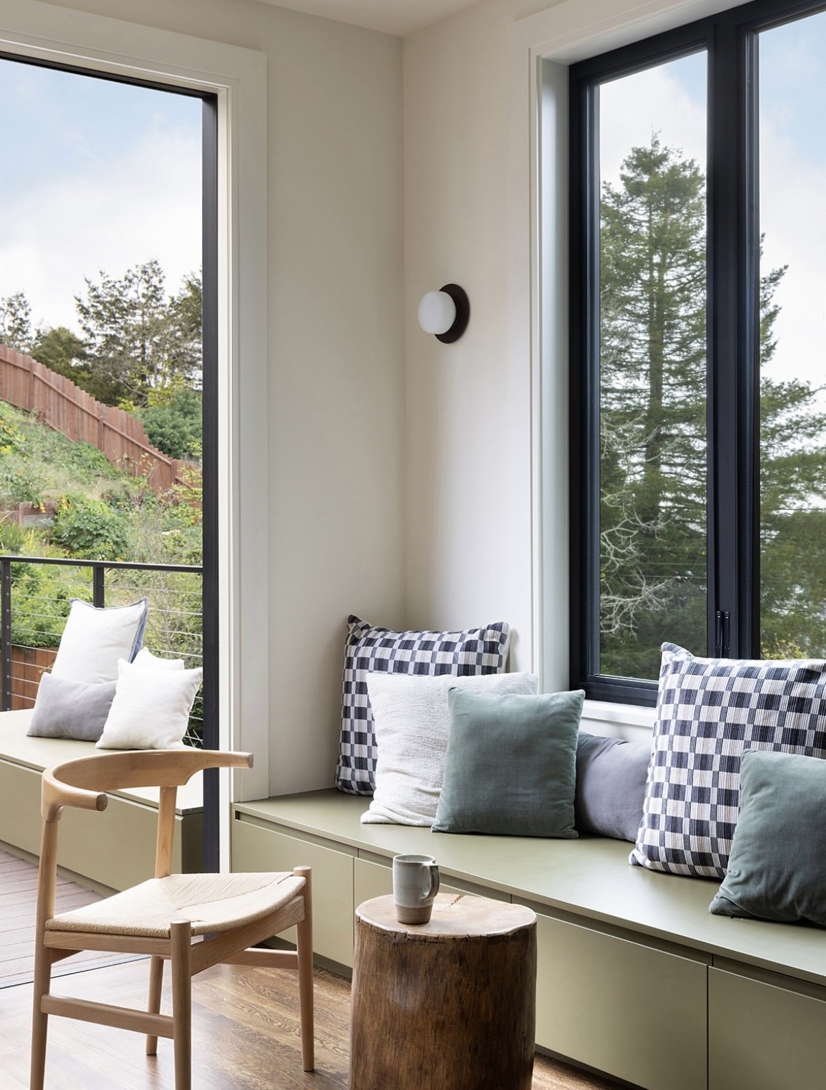 Sunlit living room corner with a built-in window seat and cushion pillows, a wooden chair, a small wooden table, and large windows showing a view of trees and a hillside.