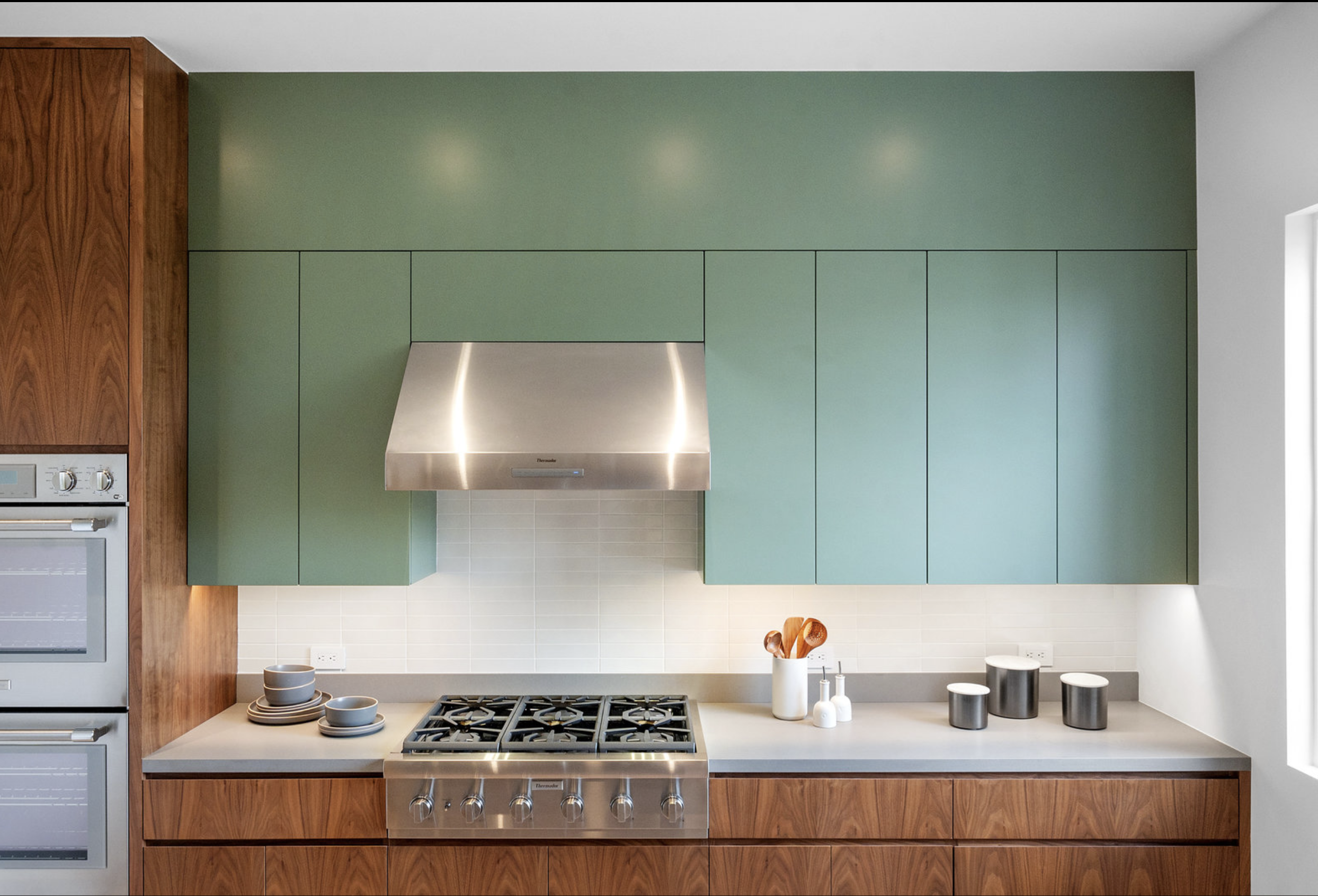 Modern kitchen with green upper cabinets, wooden lower cabinets, stainless steel range hood, cooktop, and kitchen utensils on countertop