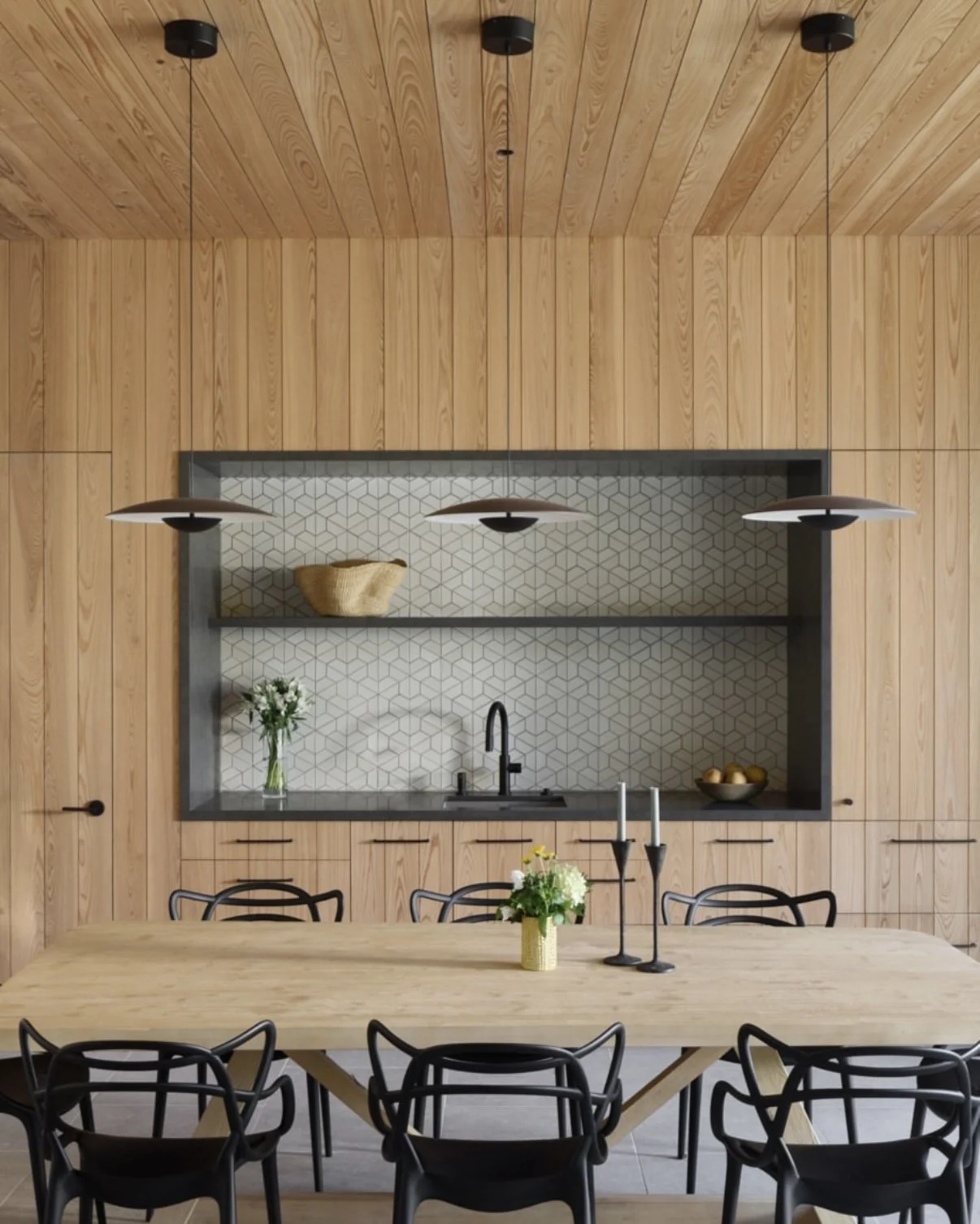 Modern kitchen with wooden panel walls, open shelving with decorative items, black sink and faucet, wooden dining table with black chairs, vase with flowers, and hanging pendant lights.