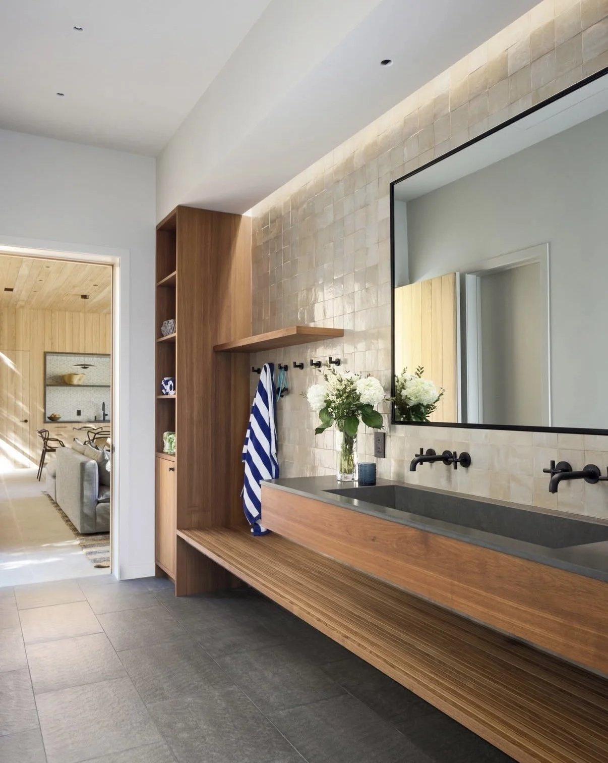Modern bathroom with a wooden vanity, large mirror, white flowers in a glass vase, navy blue candle, and beige tile wall, with a view into a living room.