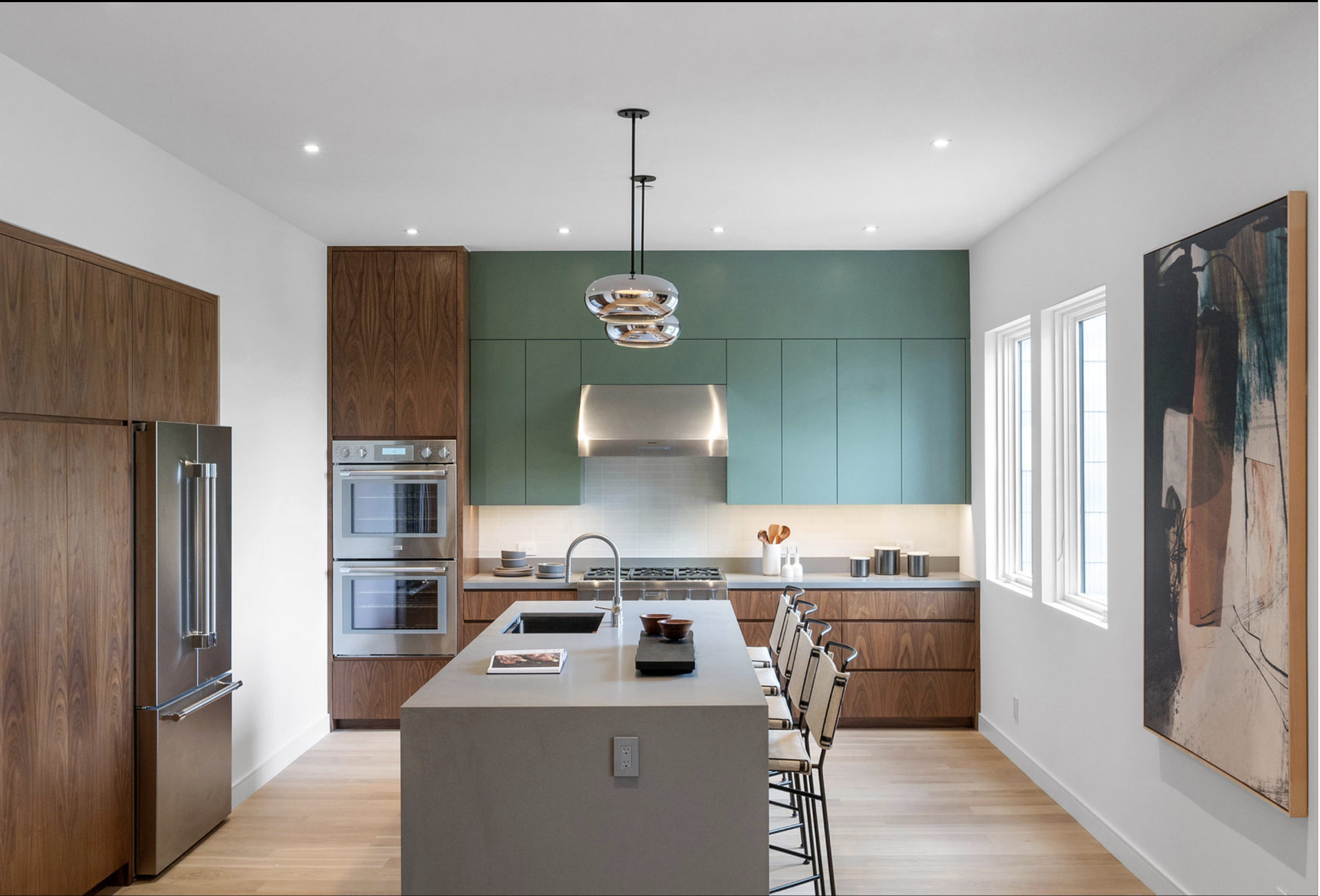 Modern kitchen with green upper cabinets, wood lower cabinets, stainless steel appliances, a central island with beige countertop, and artwork on the wall.