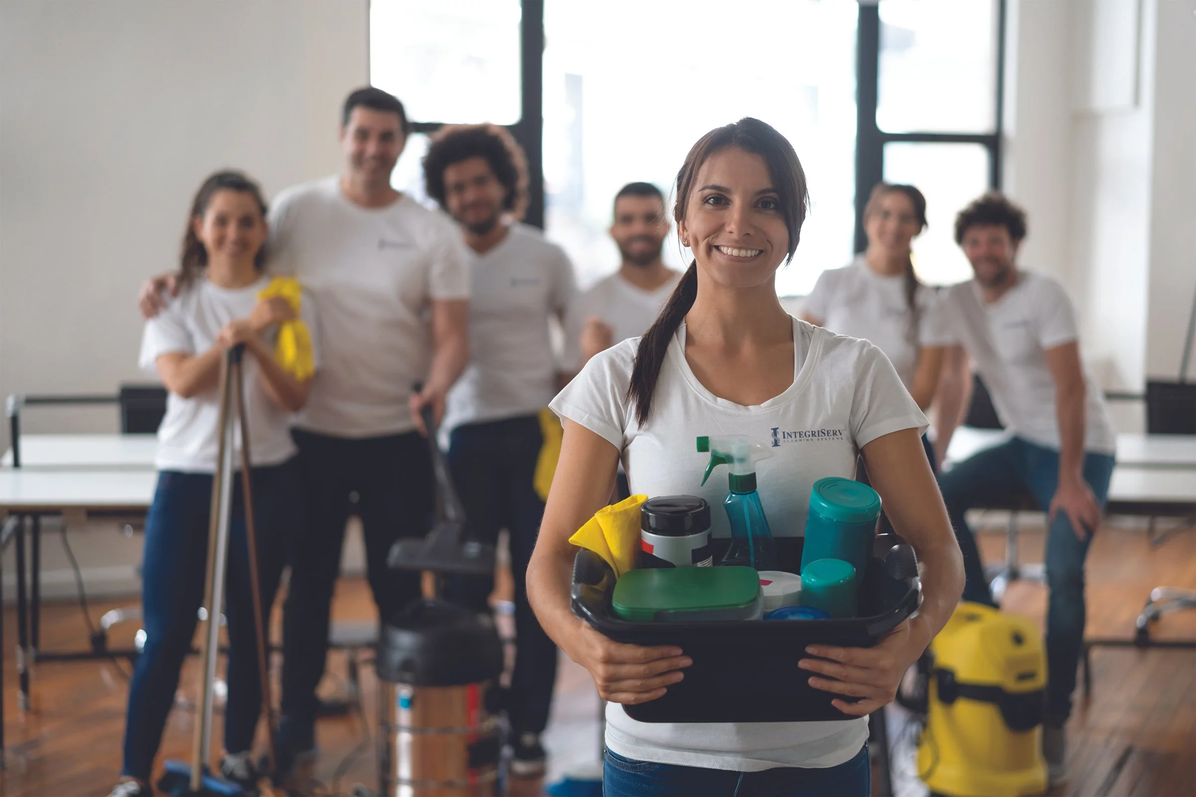 IntegriServ Cleaning Systems team member holding a cleaning caddy with supplies, with the full cleaning crew standing behind her in a bright commercial space.
