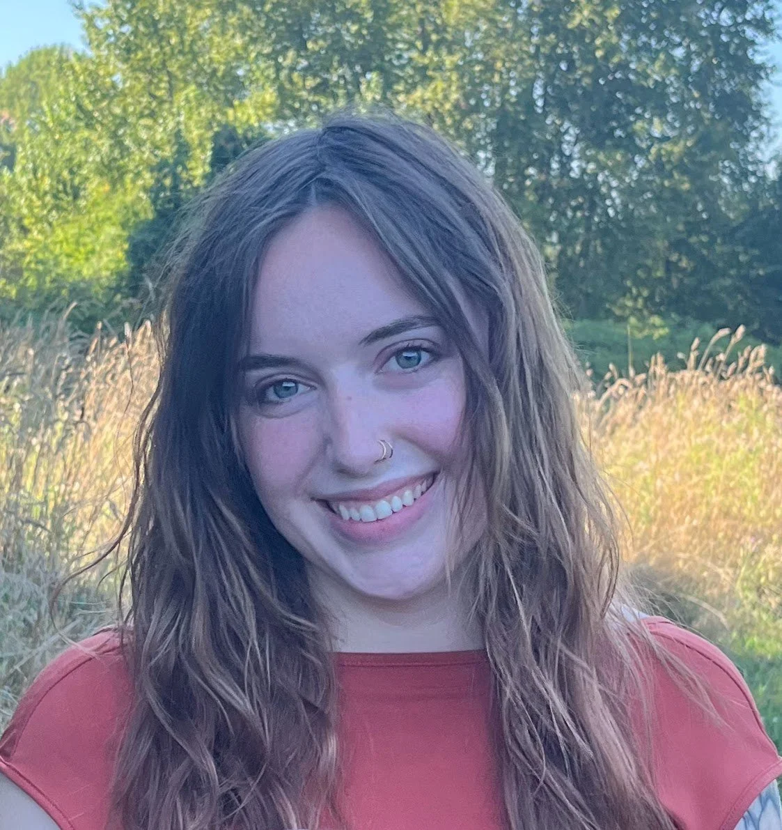 Young woman with wavy brown hair, blue eyes, and a nose ring, smiling outdoors in a field with green trees and tall grass behind her, wearing a red top.