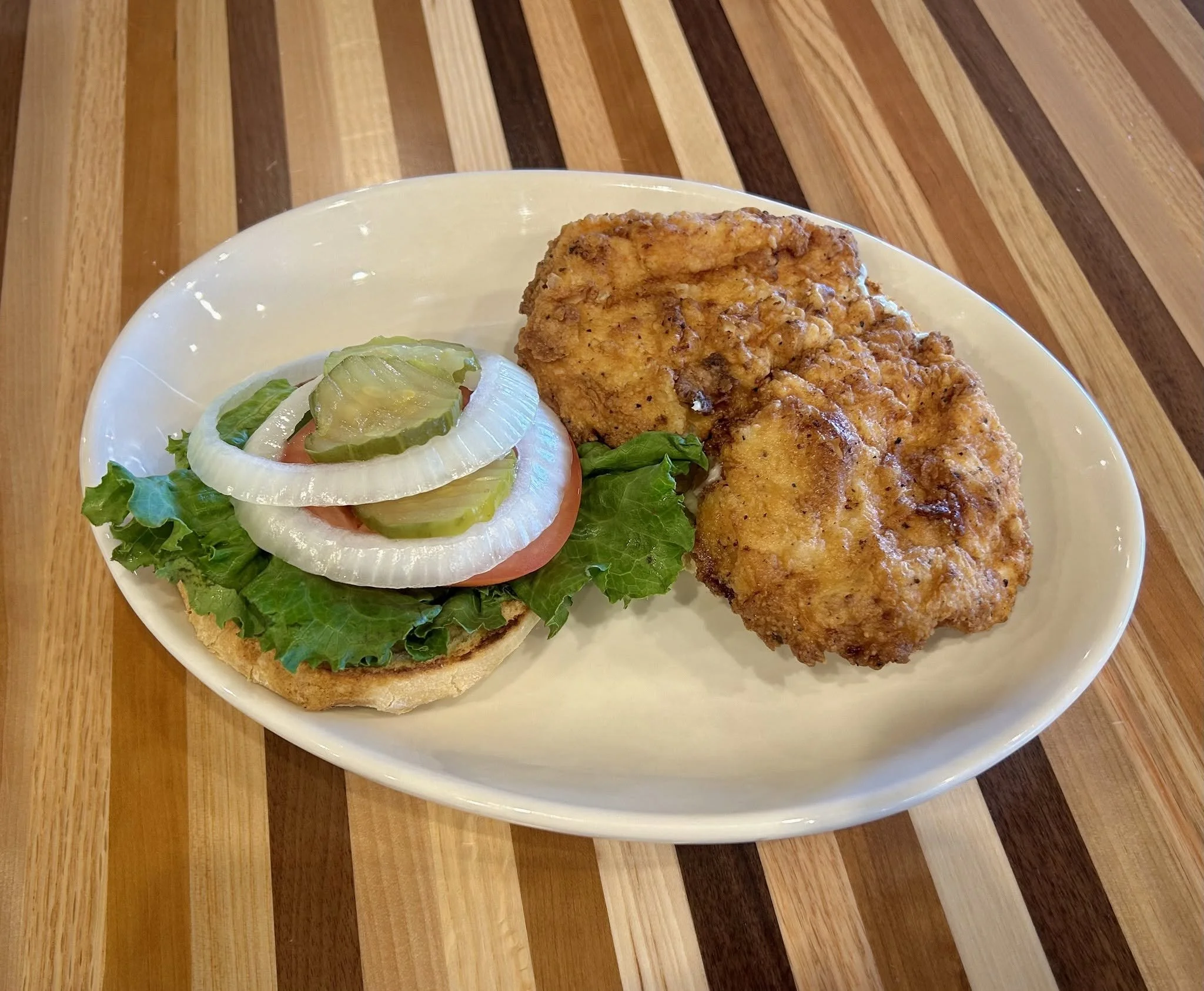 Fried fish fillet served with lettuce, tomato, onion, pickle slices, on a white, oval plate. The plate is on a wooden table.