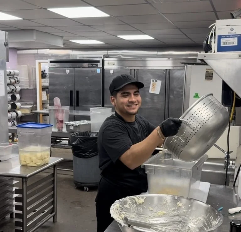 A smiling man in a black shirt and cap holding a metal strainer over a container, in a commercial kitchen with stainless steel appliances and shelves.