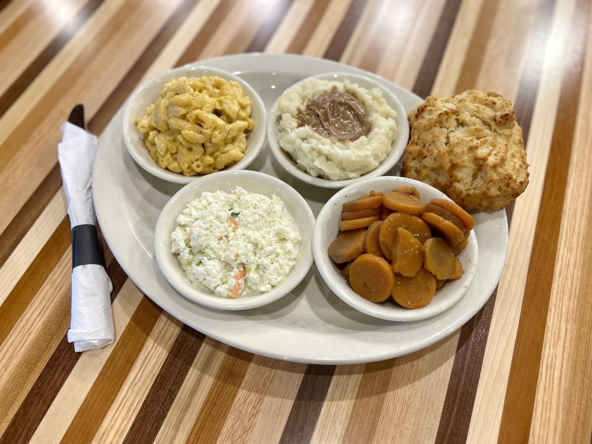 A plate with five small bowls of food, including macaroni and cheese, mashed potatoes with gravy, a scoop of potato salad, coleslaw, and a serving of cooked vegetables with sausage slices, on a wooden table with a napkin and fork wrapped in a napkin.