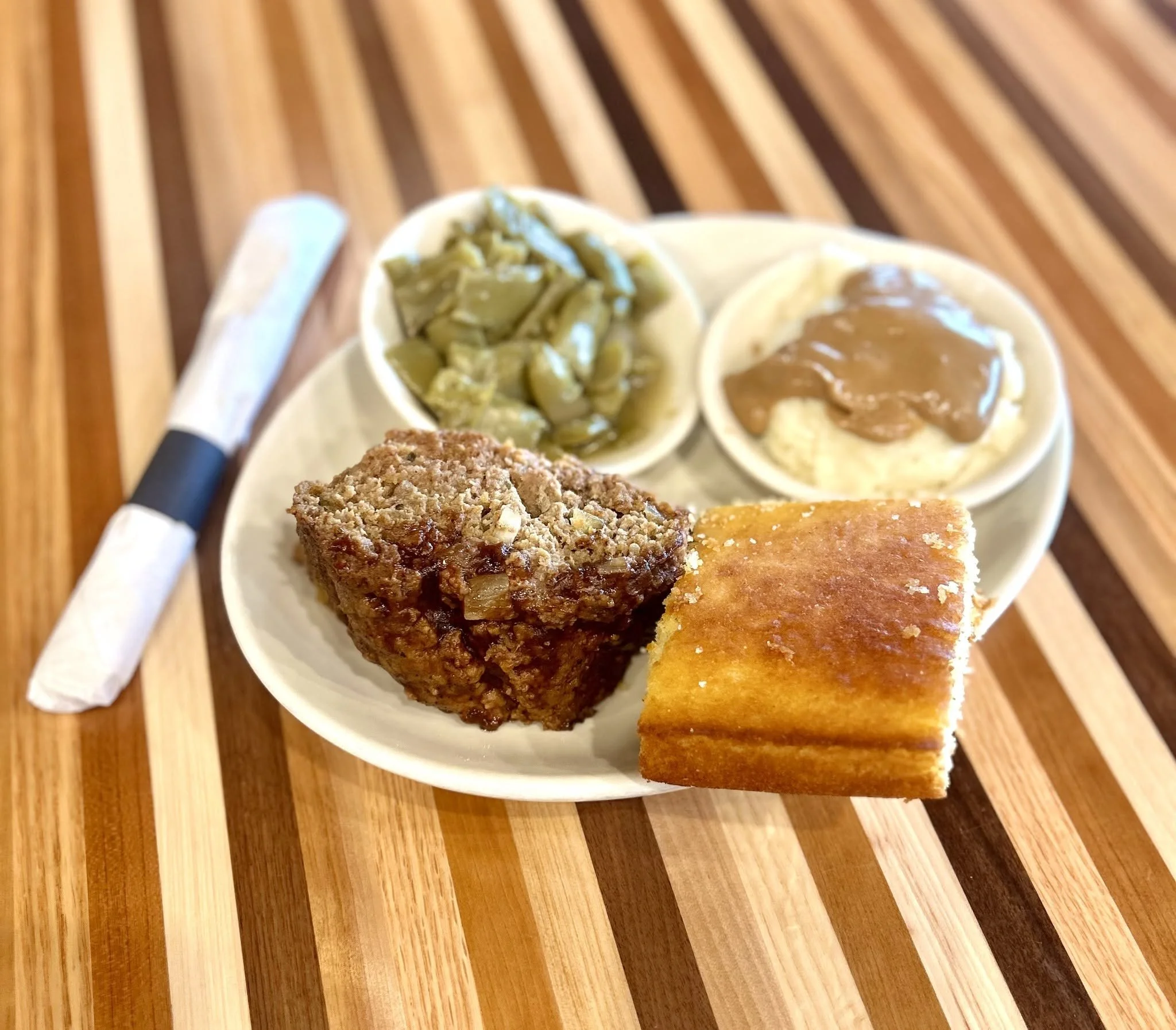 Close-up of three small bowls with different toppings on a white plate, along with pieces of fried chicken and cornbread on a wooden table.