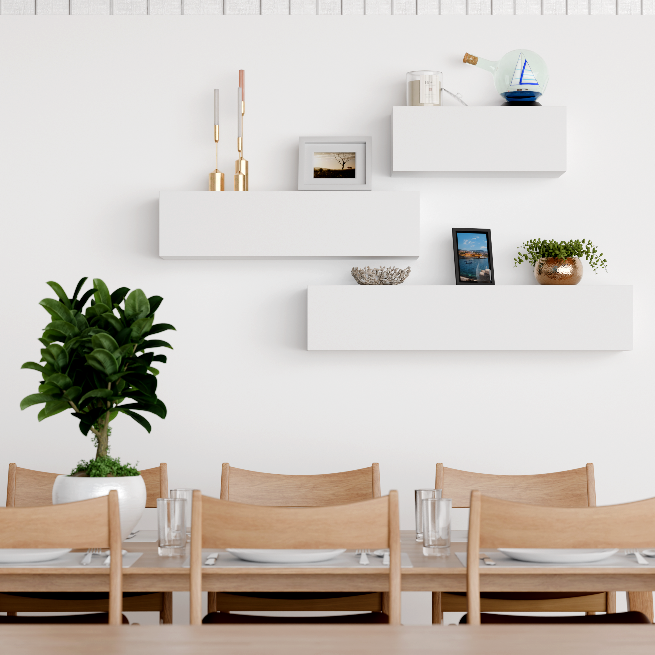 A dining room with a long wooden table set with glasses and bowls, surrounded by wooden chairs, with a white wall decorated with floating white shelves holding a framed photo, candles, a decorative glass bottle, and a small potted plant.