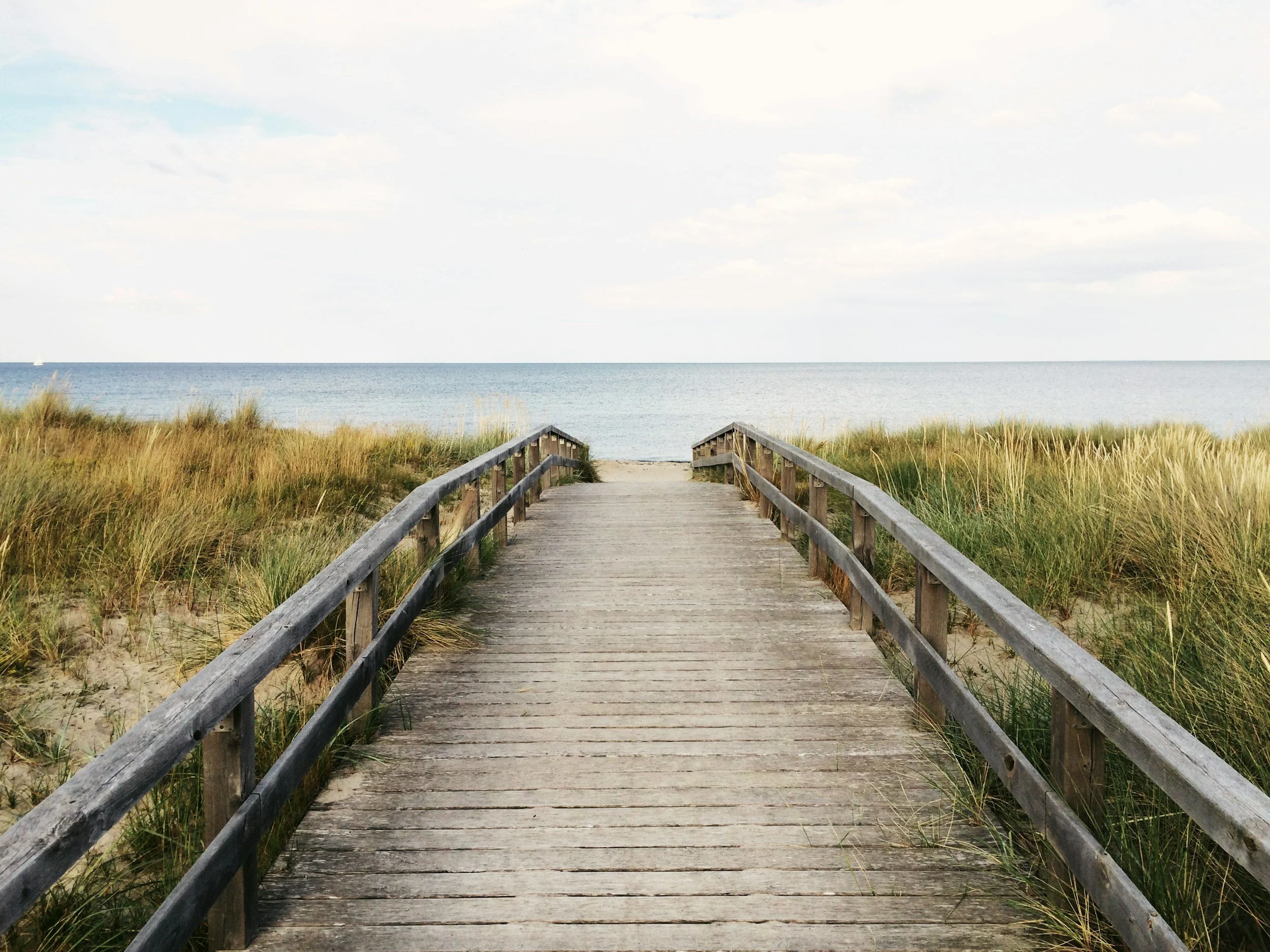 A wooden boardwalk leading towards the beach with sandy dunes and grass on either side, and the ocean in the background under a partly cloudy sky.