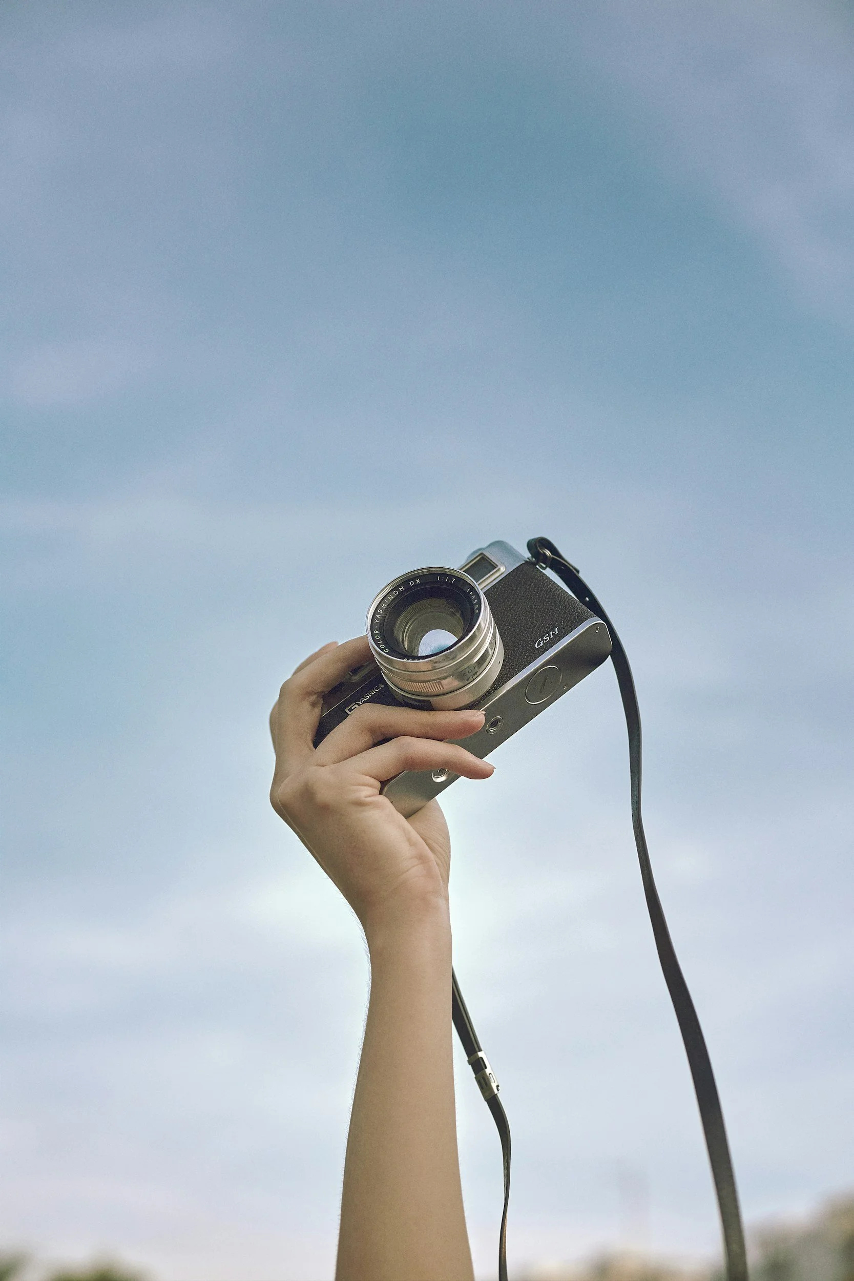A person's hand holding a vintage camera up against a bright blue sky with some clouds.
