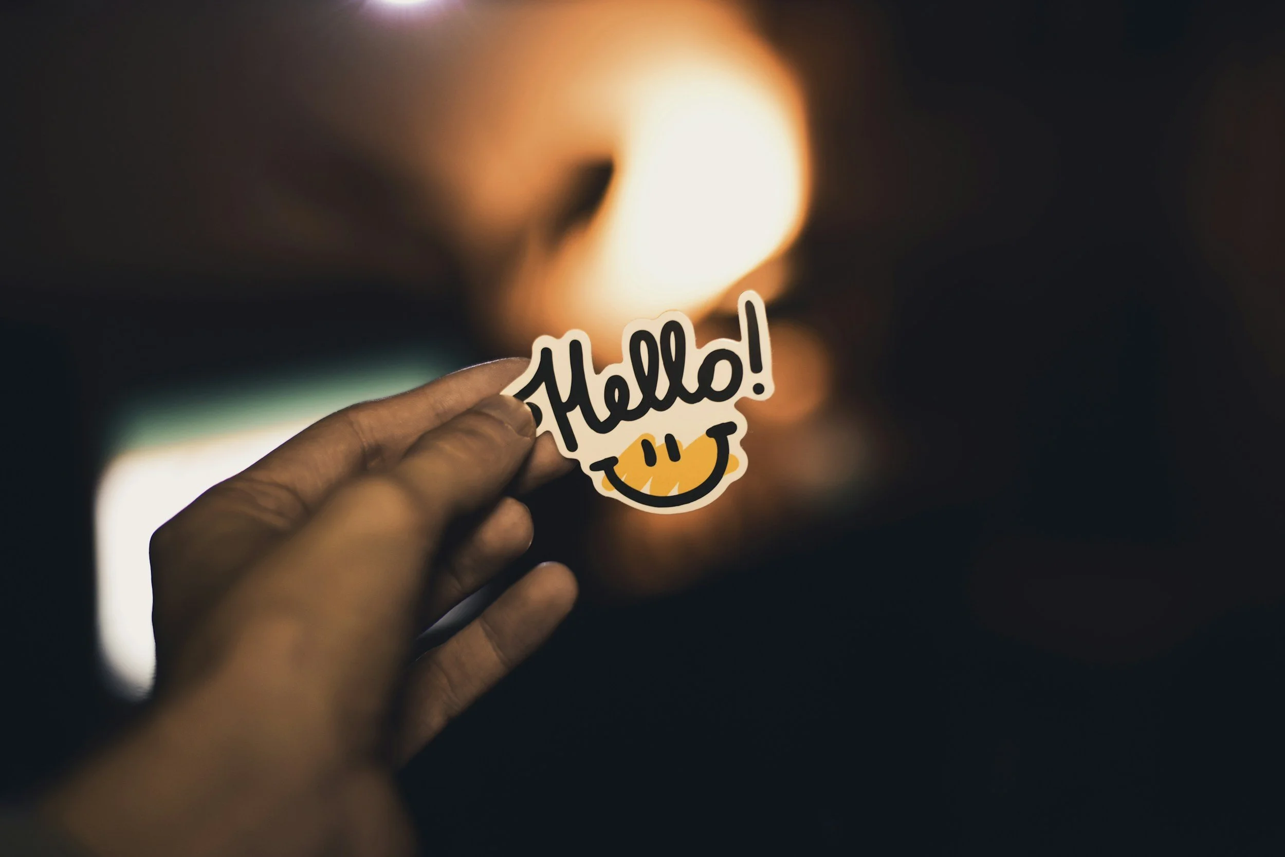 A hand holding a sticker that says 'Hello!' with a smiley face, in front of a blurred background with bright lights.