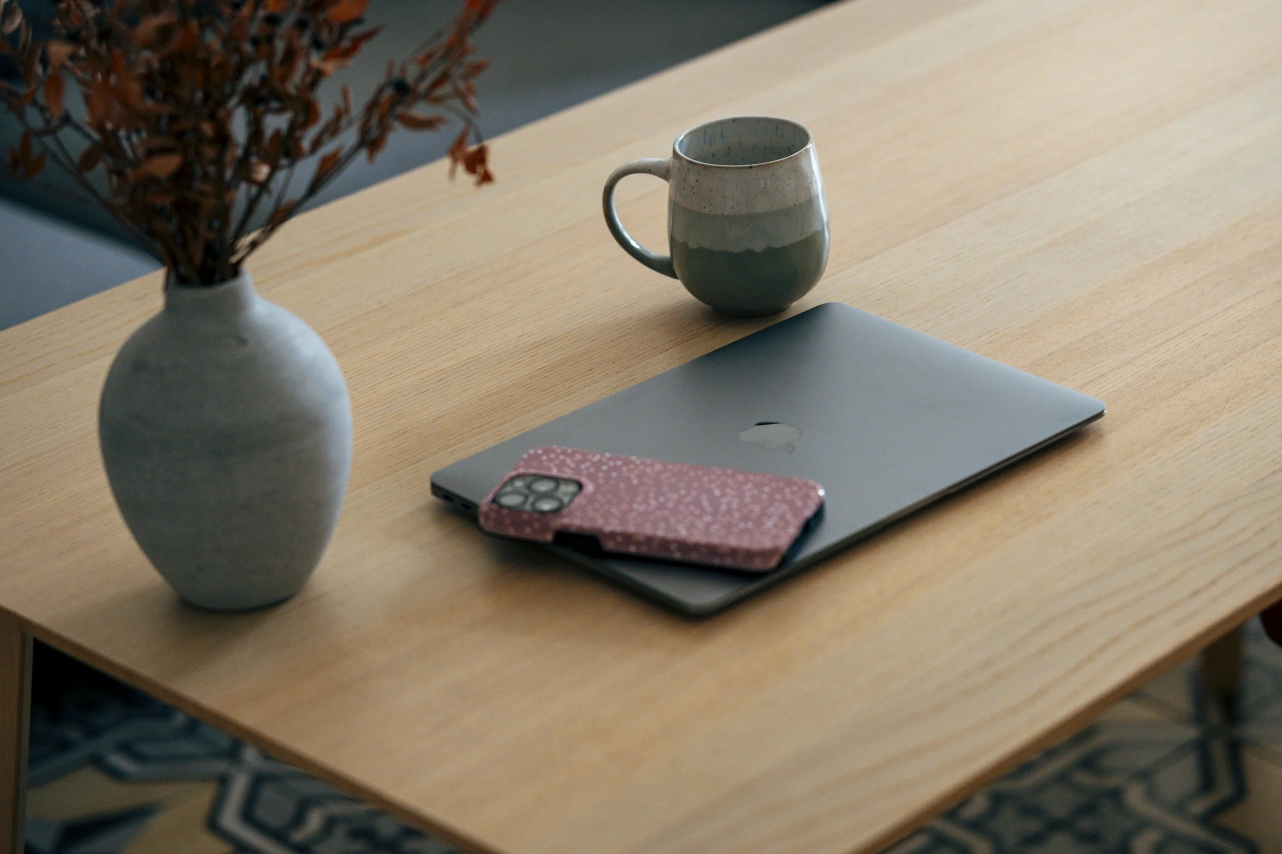 A light wood table with a white vase holding brown dried flowers, a closed silver laptop, a pink patterned phone case, and a small ceramic mug with blue and white speckled glaze. The background includes a patterned carpet.
