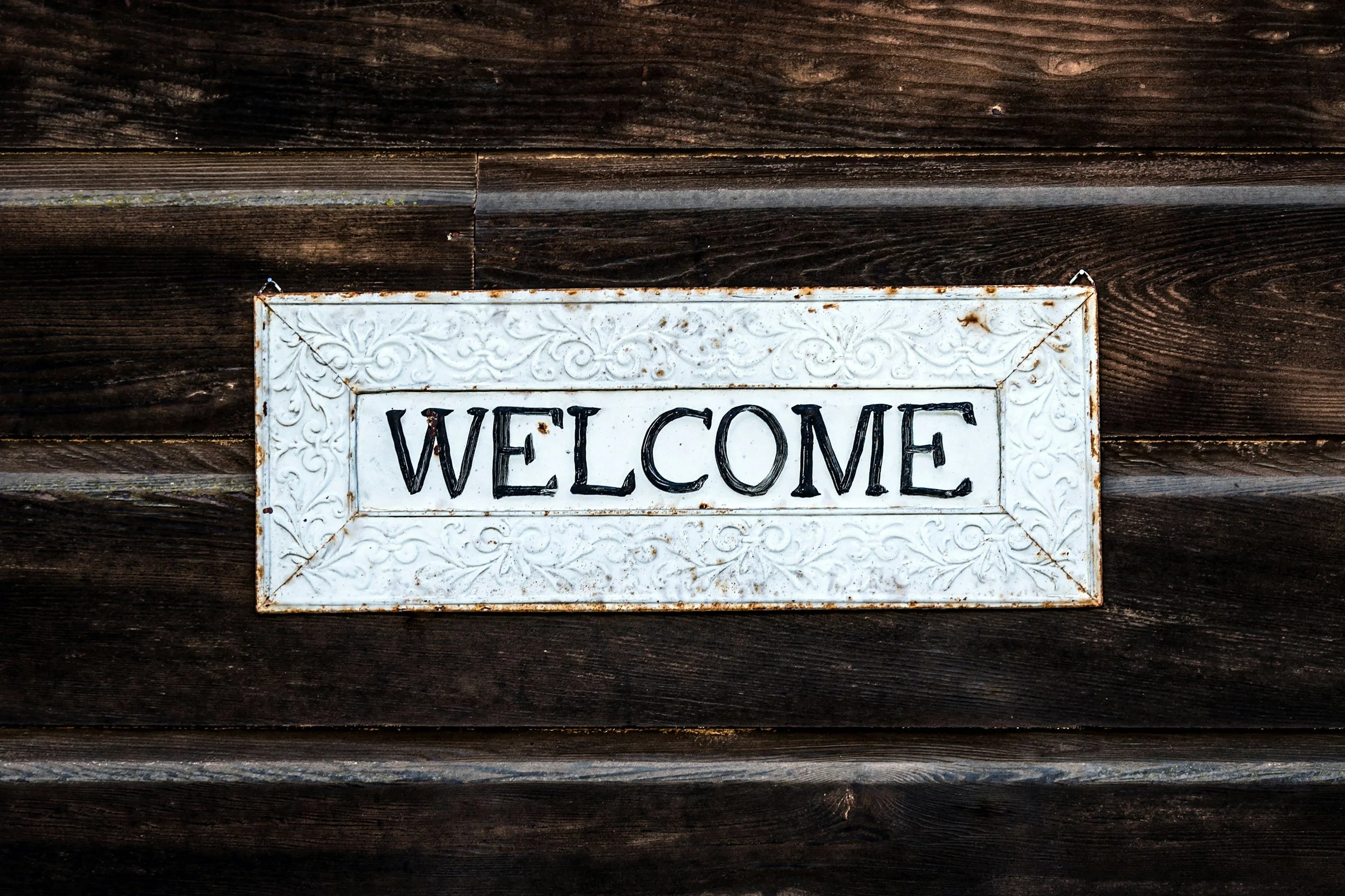 A white metal welcome sign with ornate detailing, mounted on a dark wooden wall.