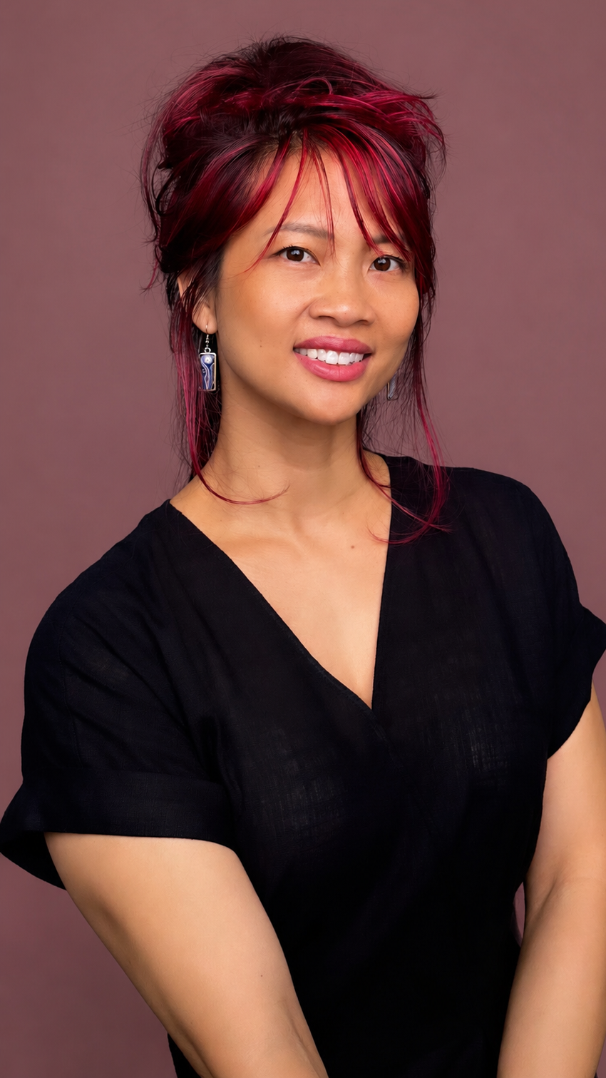A woman with short, layered, dark hair with red highlights, wearing a black top and earrings, smiling against a plain gray background.