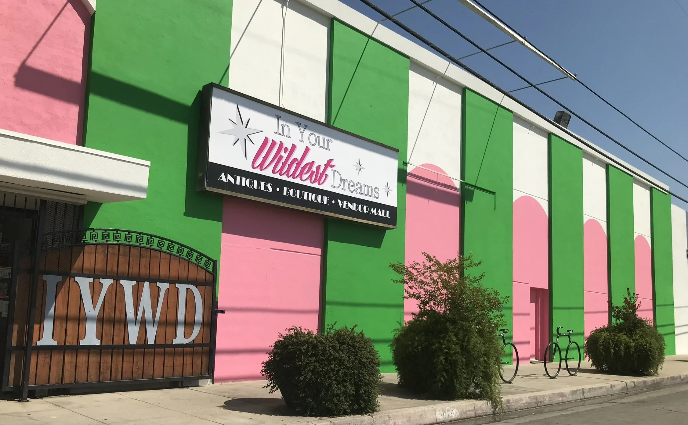 The exterior of a colorful building painted pink, green, and white, with a sign that reads 'In Your Wilderness Dreams' and tags 'Antiques, Boutique, Vendor Mall.' There is a decorative gate with 'YWD' on it, some bushes, and bike racks outside under a clear blue sky.