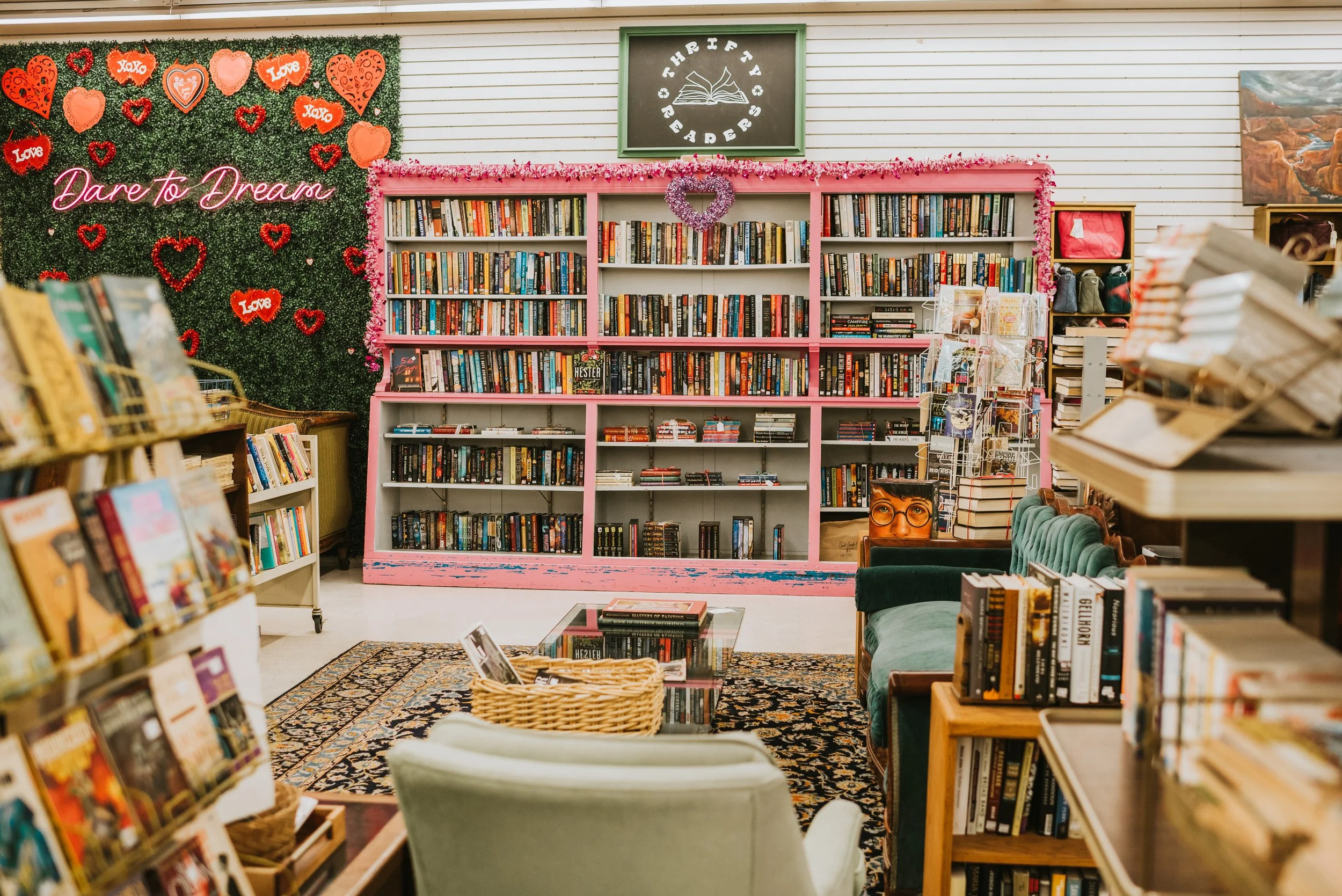 Interior of a bookstore with shelves filled with books, decorated with pink and red hearts, flowers, and the phrase 'Dare to Dream' in pink cursive lighting. There is a green velvet chair and a patterned rug on the floor.