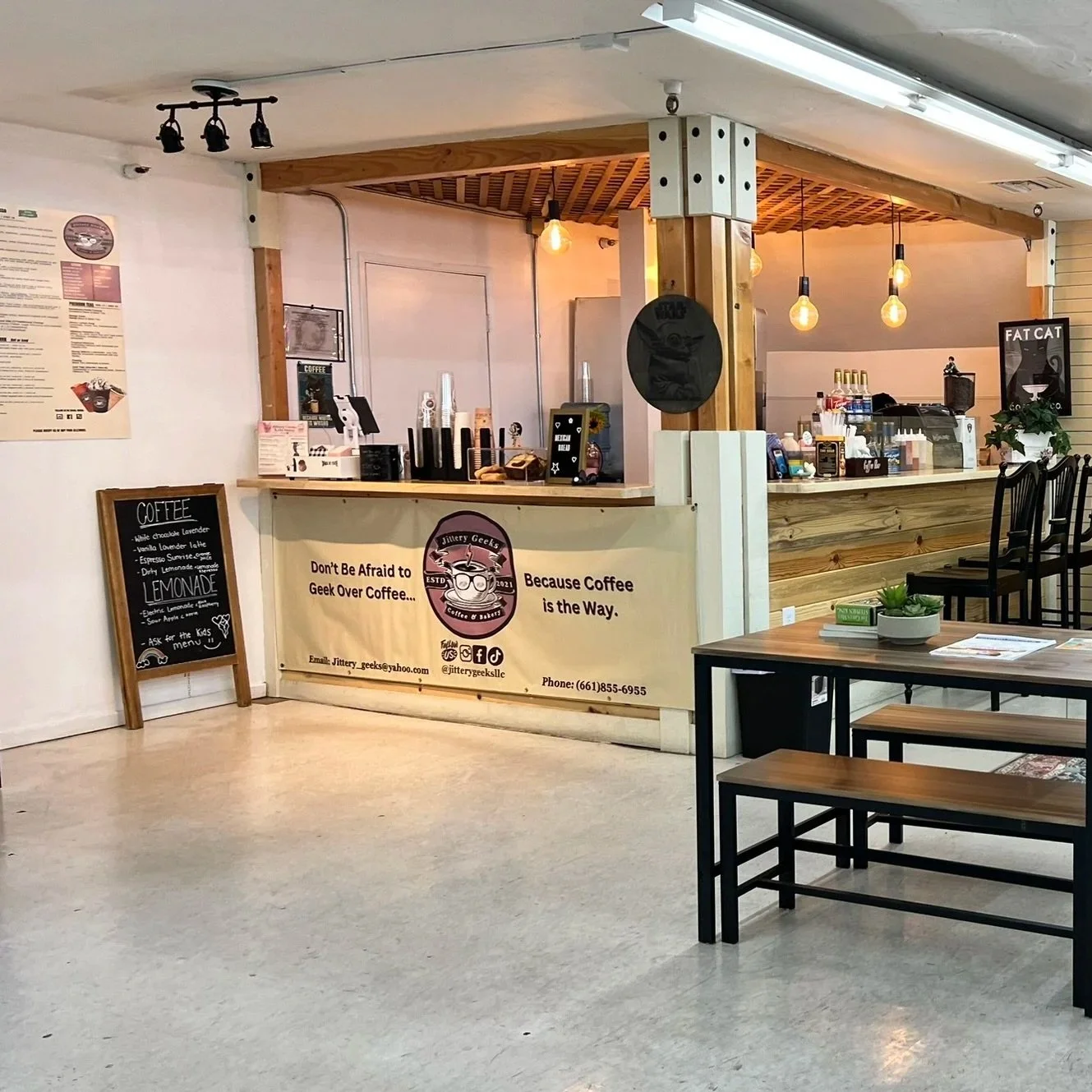 Interior of a cozy coffee shop with a counter, menu board, and seating area with tables and chairs. Decor includes wooden accents and hanging Edison bulbs.