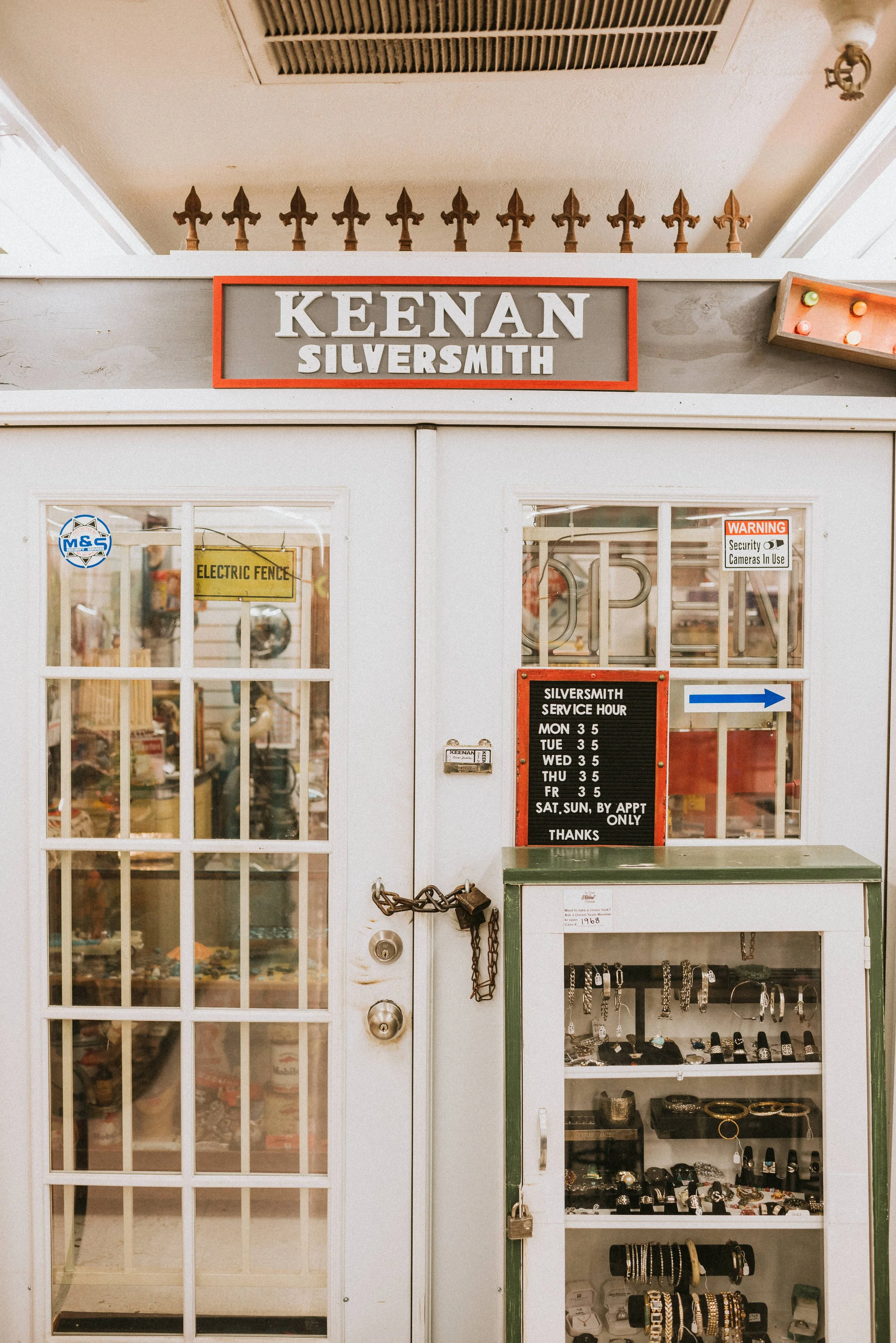 The exterior of a jewelry shop with a sign that reads 'Keenan Silversmith.' The shop has a glass door with a sign indicating service hours and a warning about security cameras, and a display case with jewelry inside.