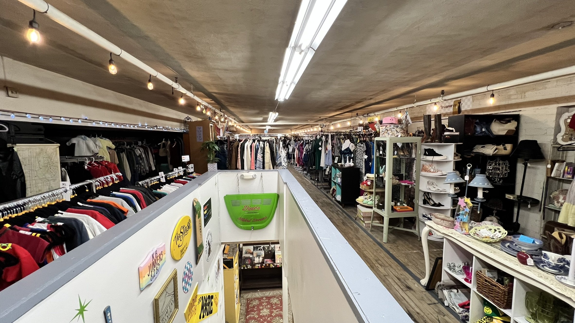 Interior view of a thrift store with clothing racks, shelves of shoes and accessories, and various household items on display, under a ceiling with exposed pipes and hanging string lights.