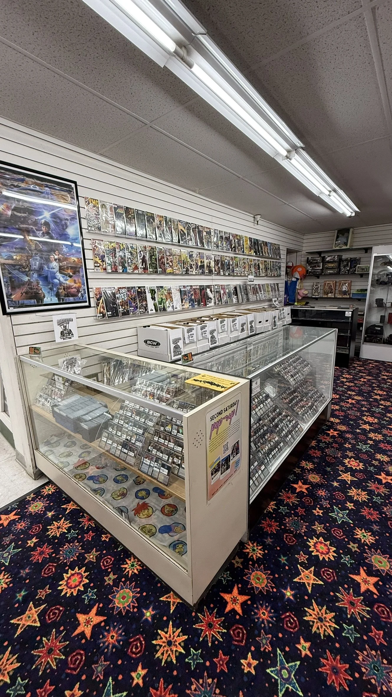 Inside a comic book store with comic books displayed on wall shelves, a glass display case filled with collectible items, and a colorful star-patterned carpet.