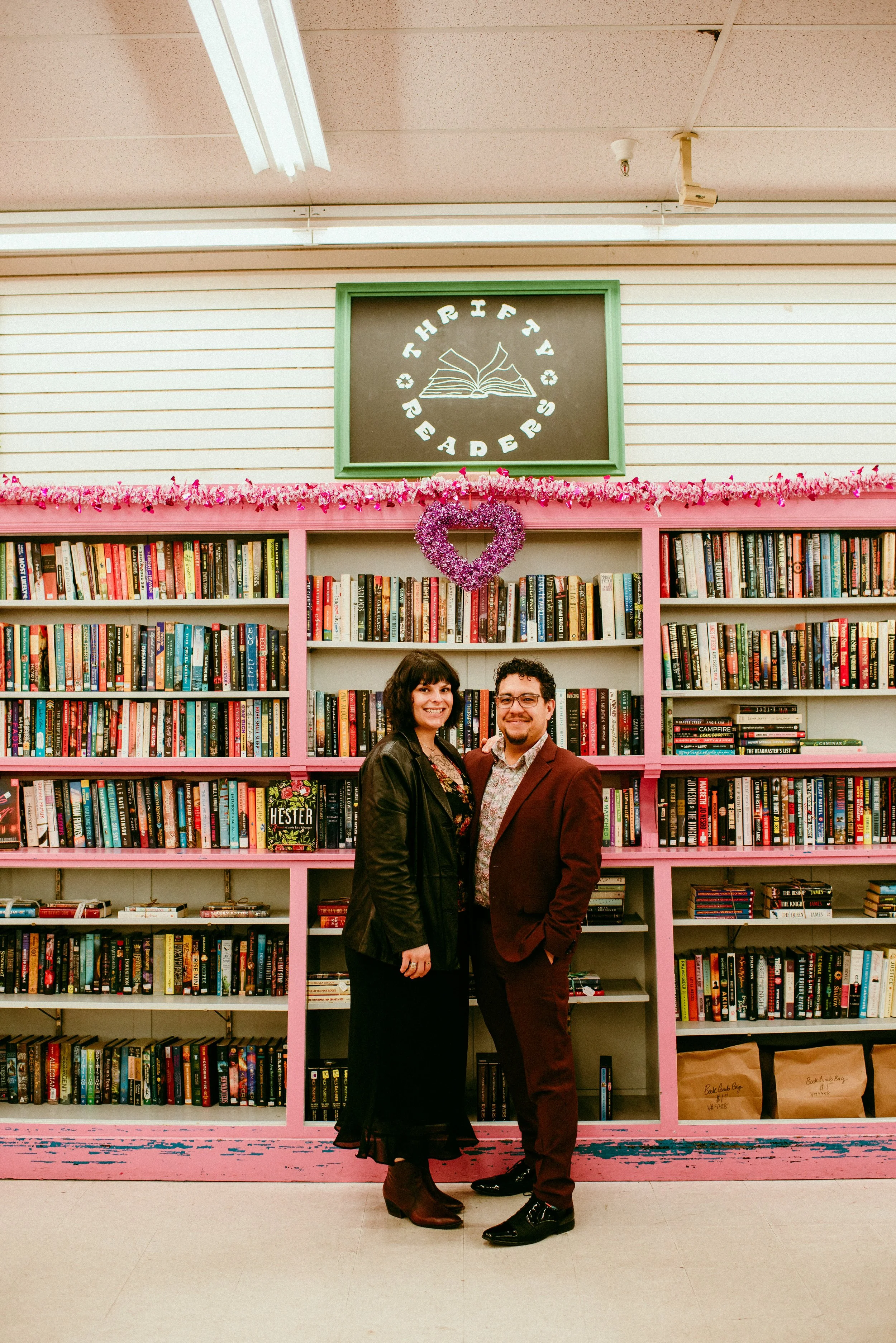 A man and woman standing in front of pink bookshelves filled with books in a bookstore. The woman has dark hair and is wearing a black leather jacket and boots. The man has curly hair and glasses, wearing a maroon suit with a floral shirt. There is a heart-shaped decoration hanging above them and a sign reading 'Third Floor Readers' with an open book illustration.