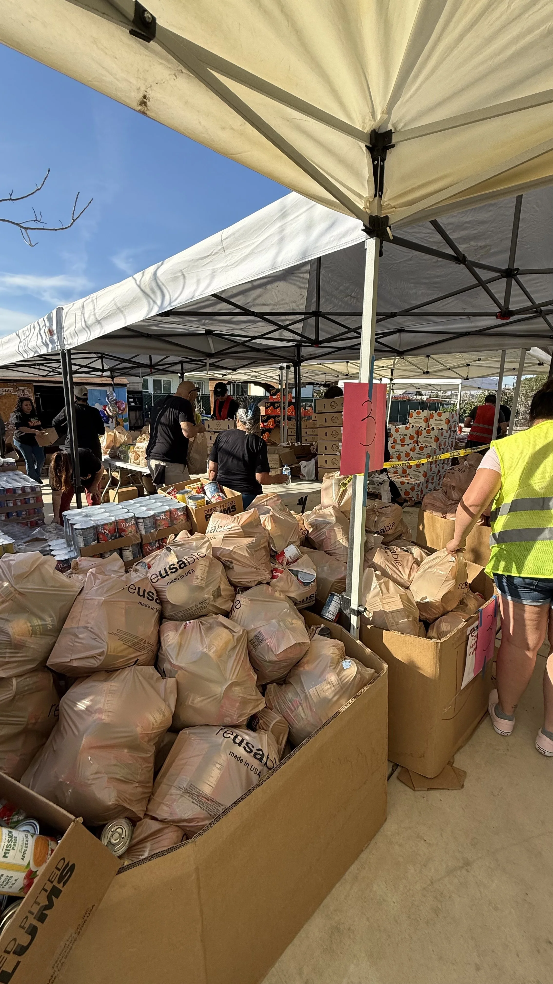 People under tents at a food distribution event with boxes of canned goods and produce.