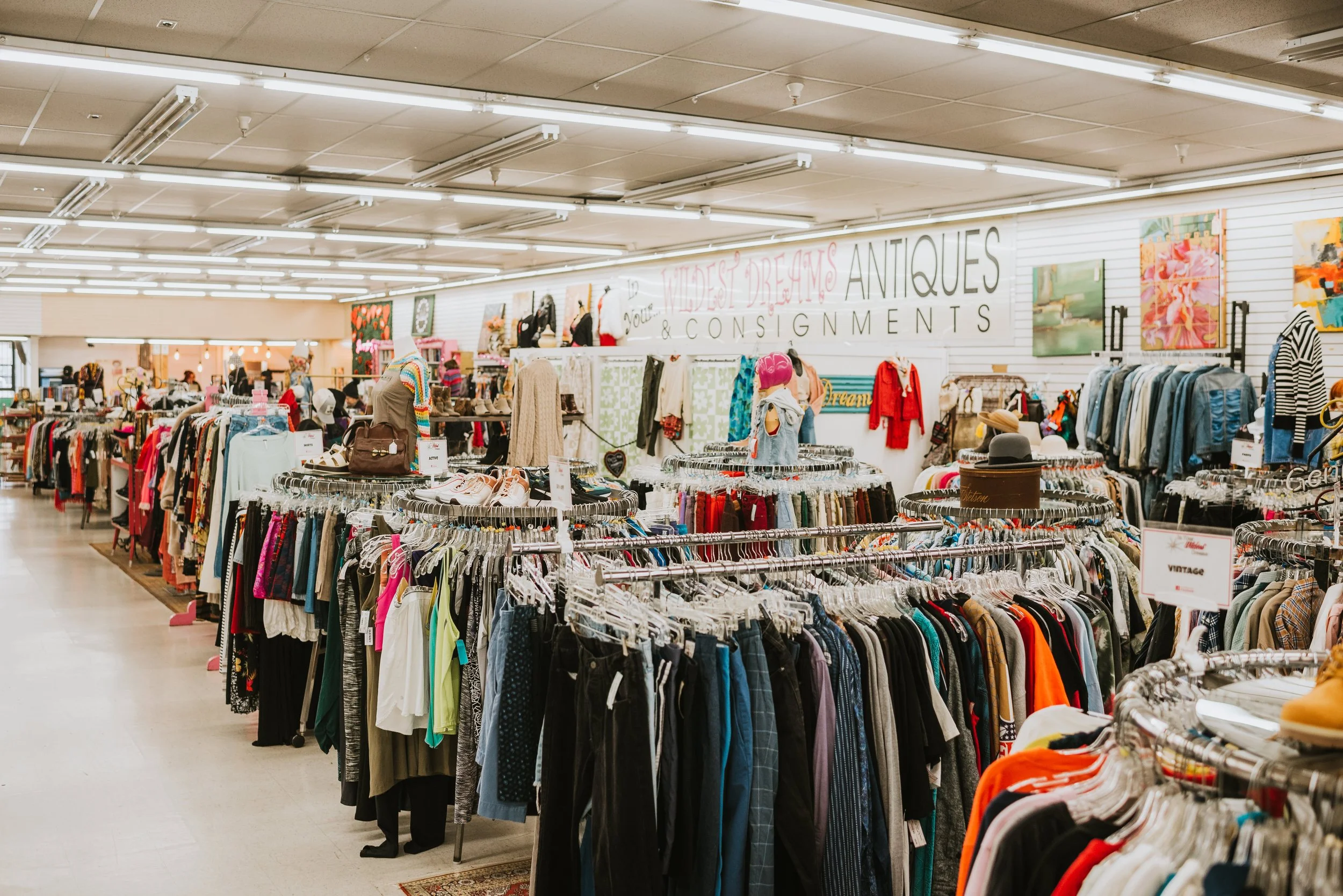Interior of a thrift store or vintage clothing shop with various racks of clothes and accessories, and a large sign reading 'Mildest Dreams Antiques & Consignments' in the background.