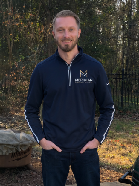 A man with a beard and light hair standing outdoors with trees in the background, wearing a navy blue Meridan Physicians Group quarter zip pullover with white accents and hands in his pockets, smiling at the camera.
