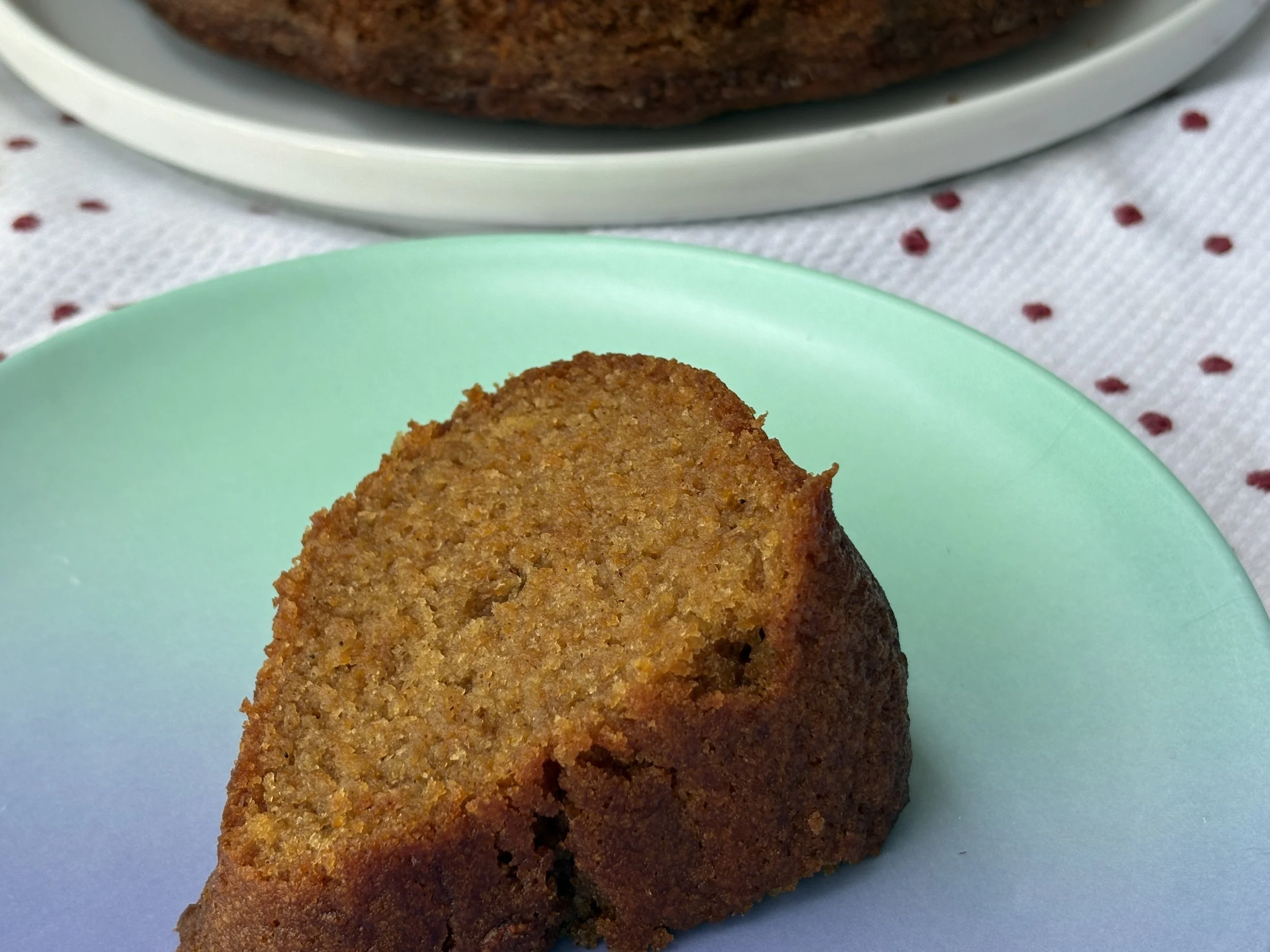 A slice of Gluten Free Carrot Cake on a green plate, with a white plate holding a bundt carrot cake in the background on a white tablecloth with red polka dots.