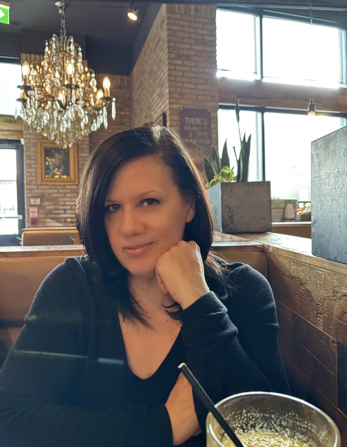 A woman with black hair wearing a black top, sitting at a restaurant table, resting her chin on her hand, with a drink in front of her. The restaurant has brick walls, large windows, a chandelier, and plants in the background.