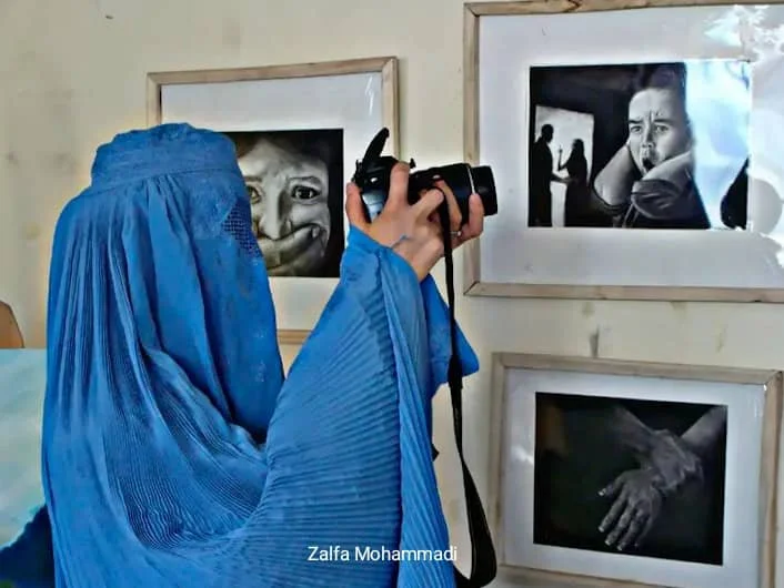 A woman in a blue headscarf and dress is taking a photograph of black and white portrait photographs on the wall, including a child's face, a woman crying, and an outstretched hand.