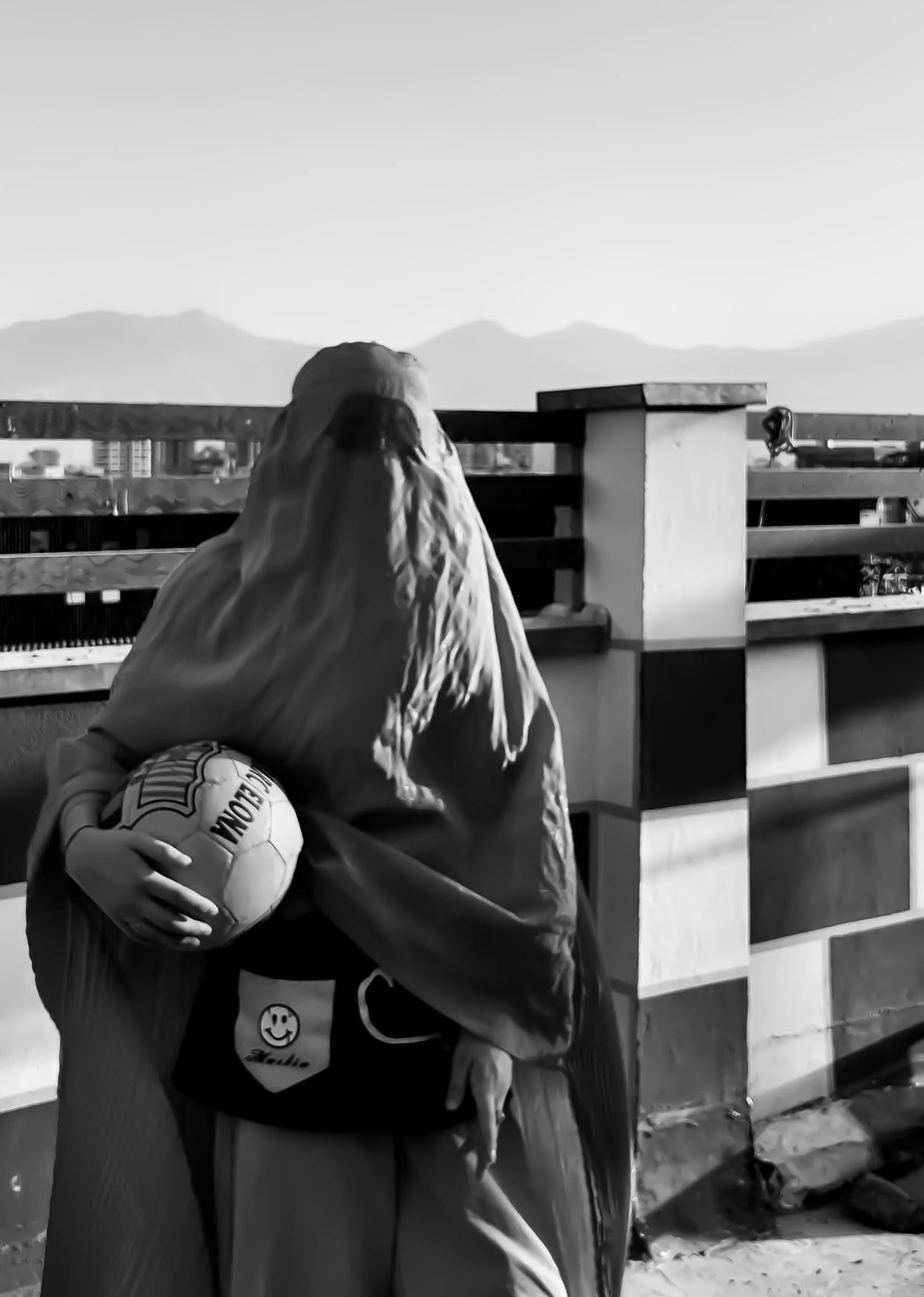 A woman with long hair sitting on a balcony, holding a soccer ball, with a cityscape and mountains in the background.