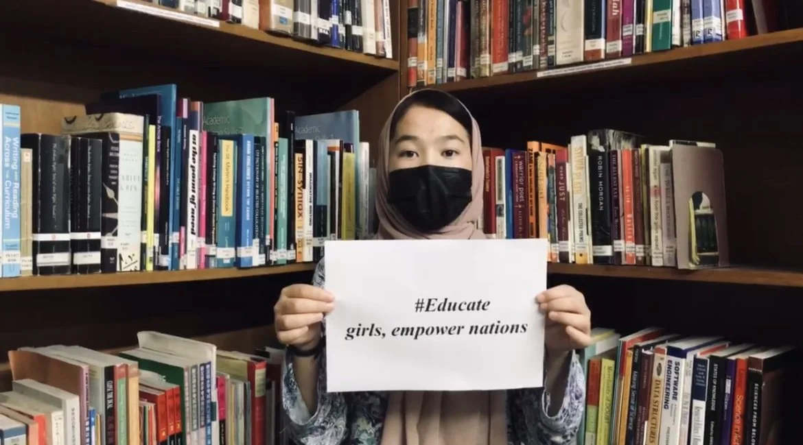 A girl wearing a headscarf and a face mask holding a sign that reads '#Educate girls, empower nations' in front of a bookshelf filled with books.