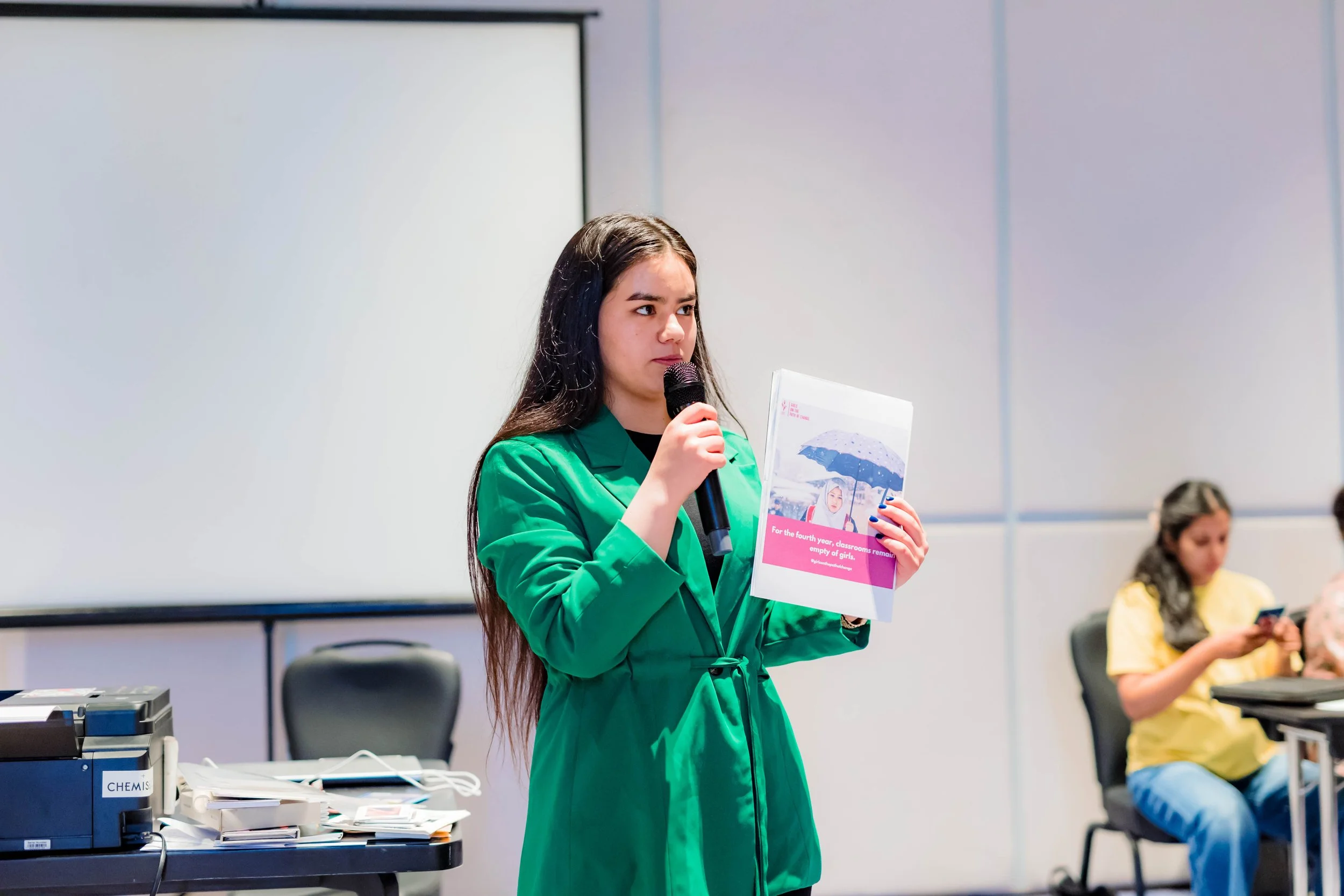 A young woman with long dark hair wearing a green blazer is speaking into a microphone while holding a booklet with a photo of children holding umbrellas. She appears to be presenting or speaking at an event or meeting, with two women seated in the background.