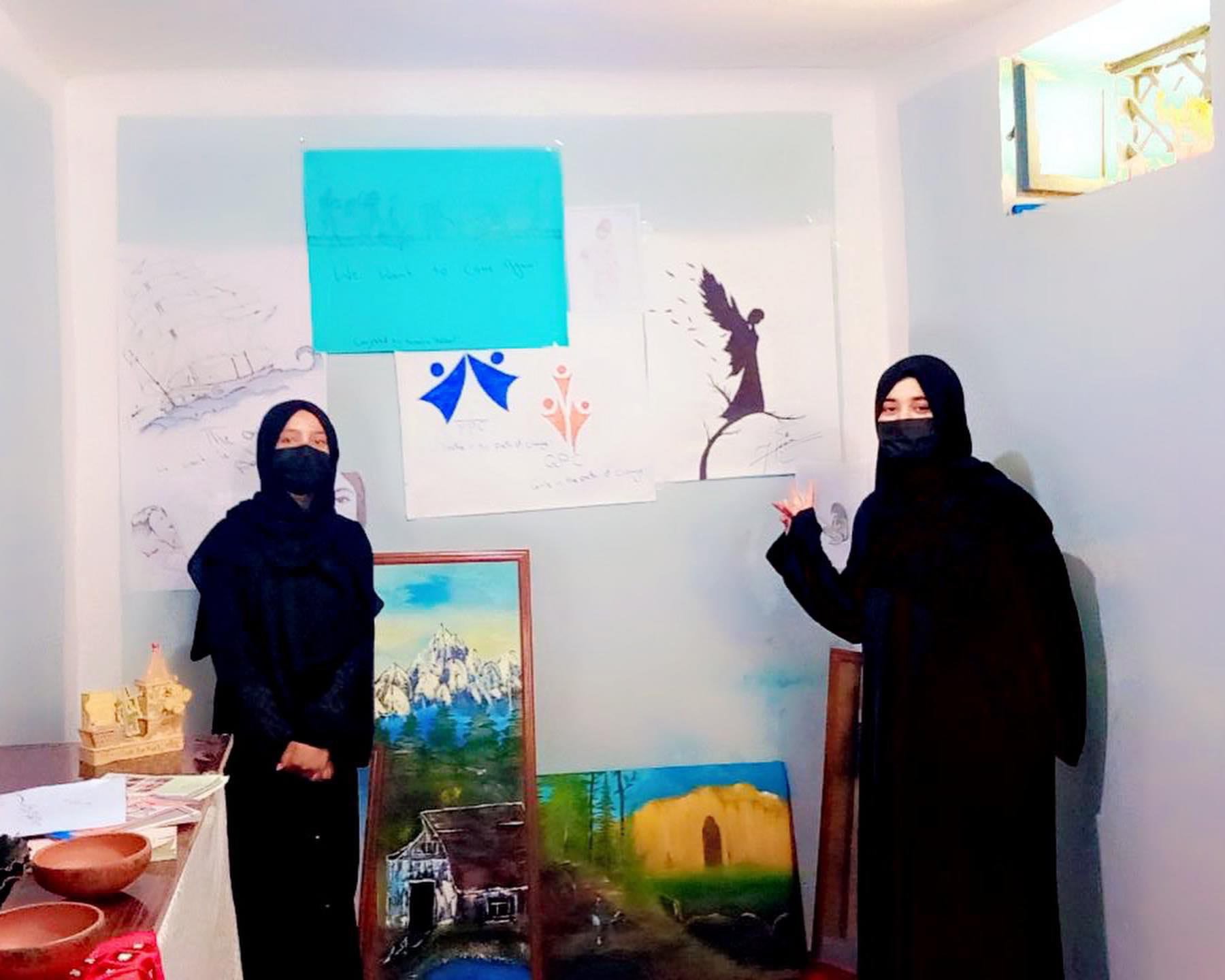 Women in black hijabs and masks stand in front of painted and drawn artworks on a wall and display easels inside an art exhibition with various paintings, including landscapes and abstract designs.